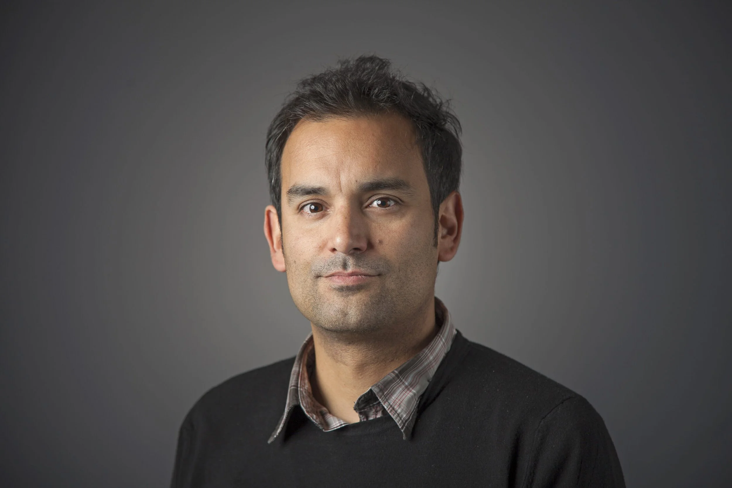 Retrato de un hombre con cabello oscuro y expresión neutral, vistiendo un suéter oscuro y camisa a cuadros, con fondo gris.