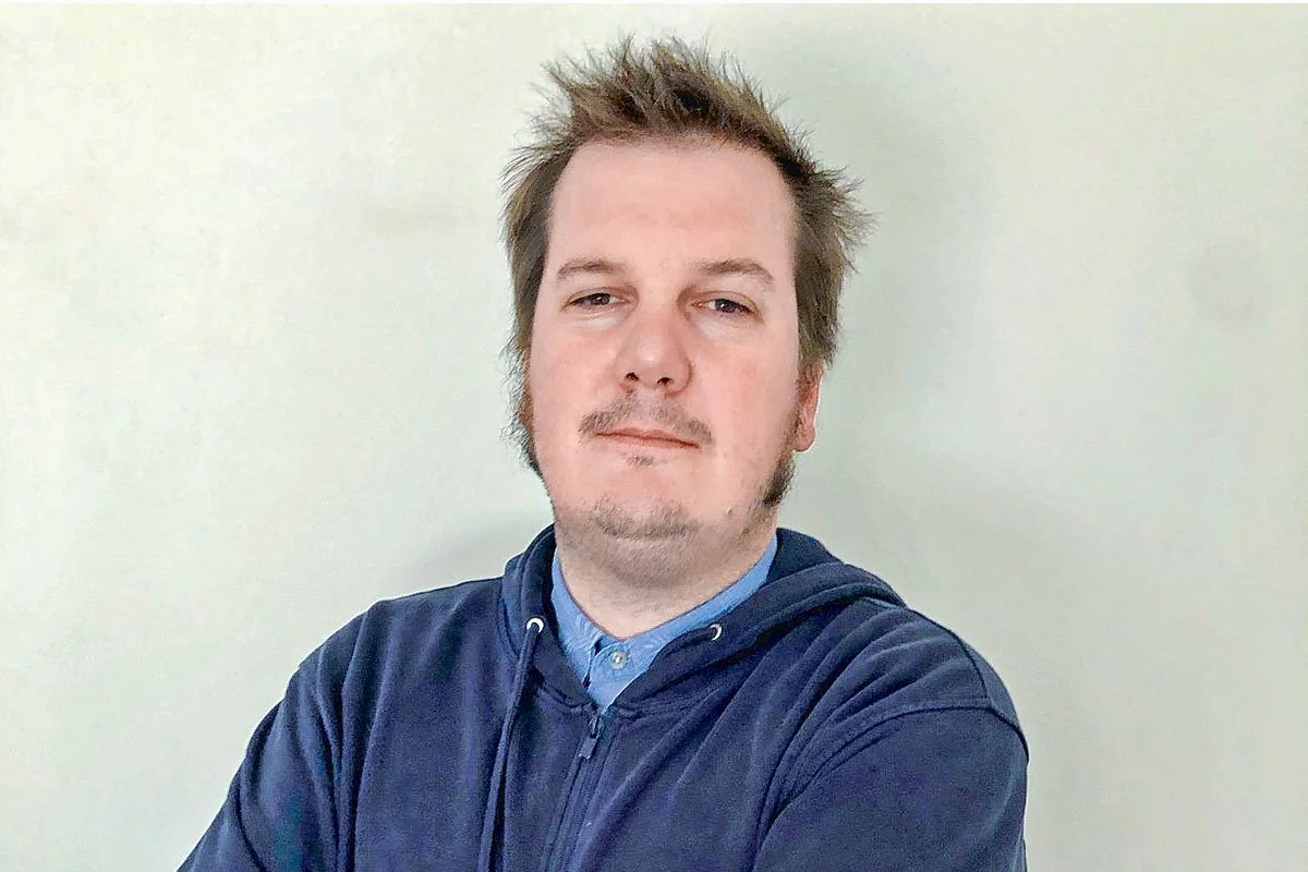 Persona con cabello castaño y barba corta, vistiendo una sudadera con capucha azul, frente a un fondo claro.