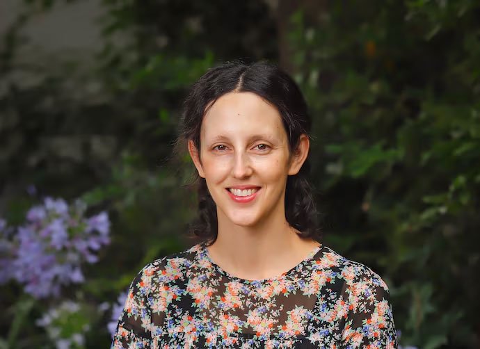 Persona con cabello oscuro y suelto, vistiendo una blusa de flores, sonriendo frente a un fondo de vegetación.