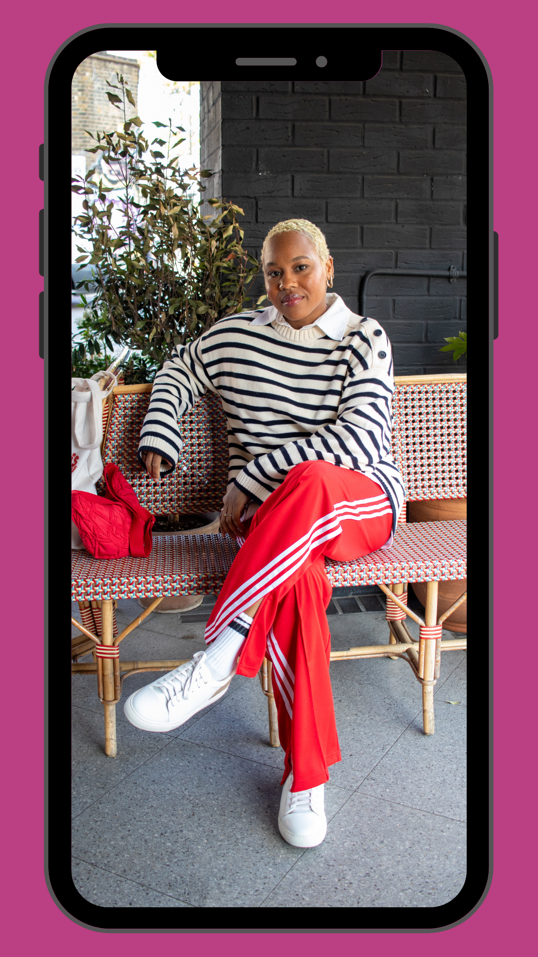 A woman with short blonde hair, wearing a white and navy striped sweater, red athletic pants with white stripes, and white sneakers, sitting on a colorful woven bench outside near a brick wall and greenery.