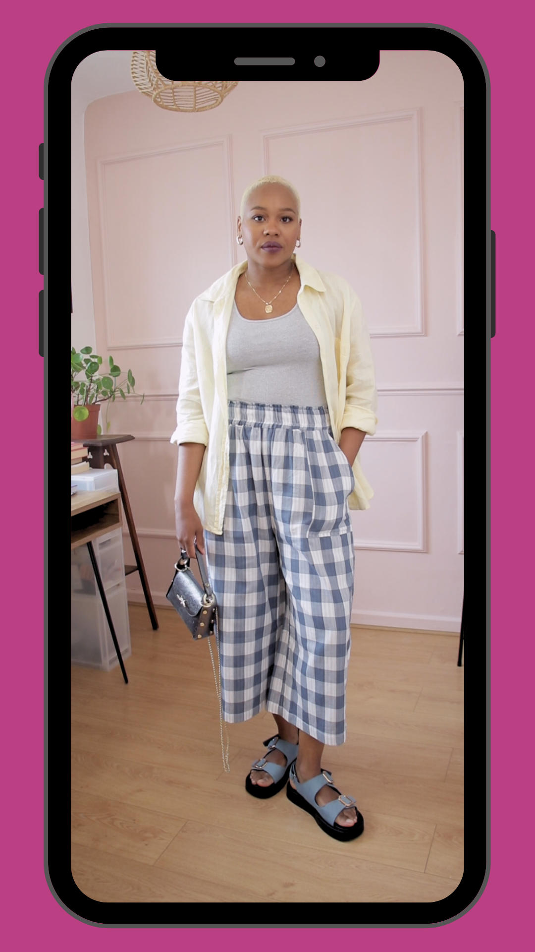 Woman with a shaved blonde hairstyle wearing a light yellow jacket over a gray tank top, checkered wide-leg pants, and blue platform sandals, standing indoors with pink walls and wood flooring, holding a small purse.