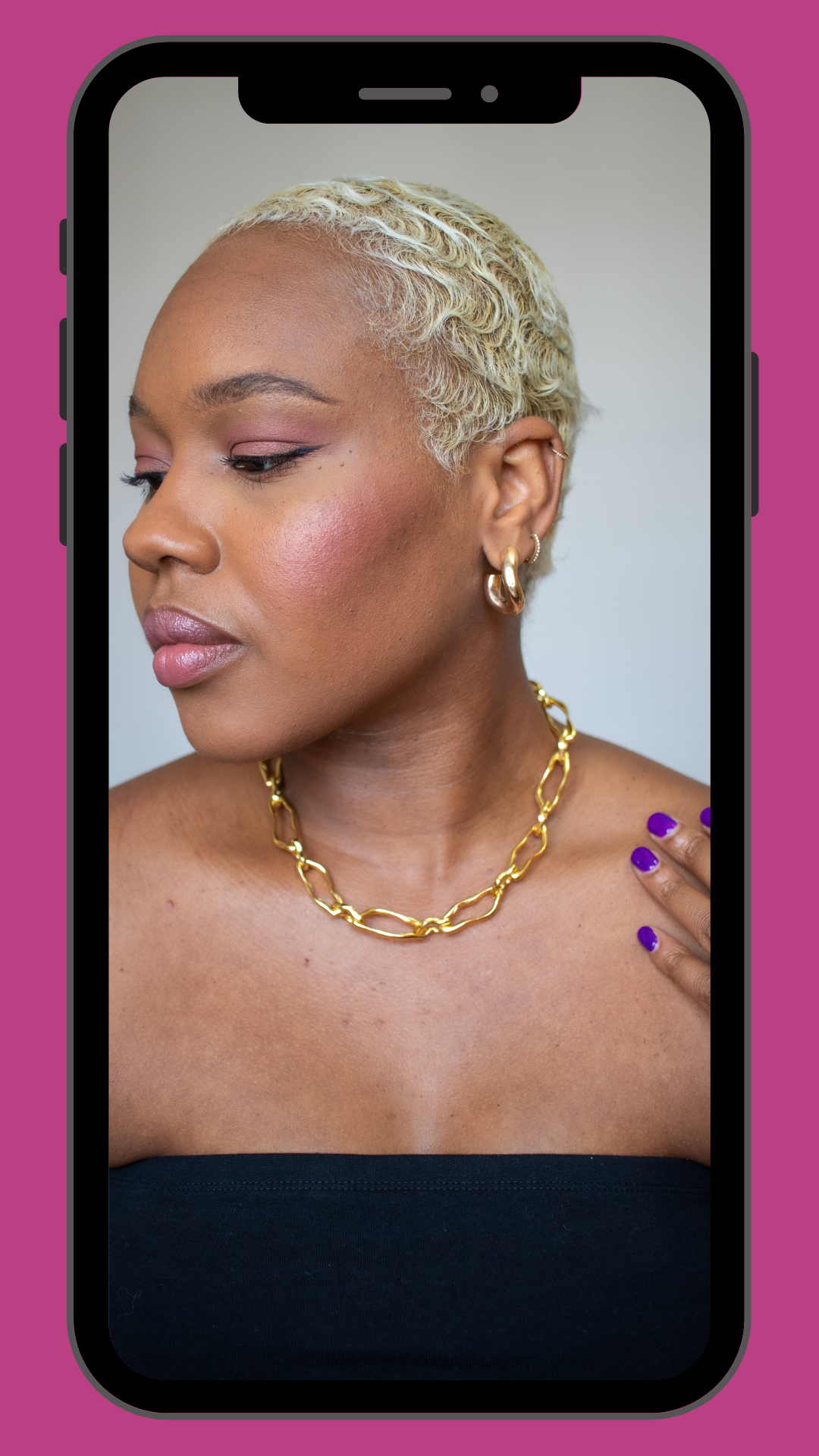 A woman with short, platinum blonde hair and medium brown skin wearing gold hoop earrings and a gold chain necklace, posing with her eyes closed and her left hand on her shoulder, against a light background.