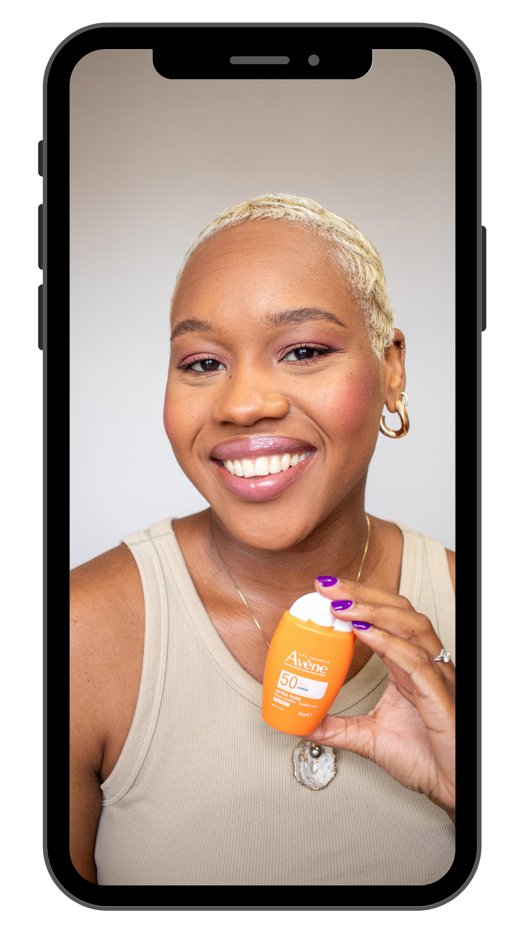 A woman with short blonde hair smiling and holding a bottle of Aveeno sunscreen lotion with SPF 50.
