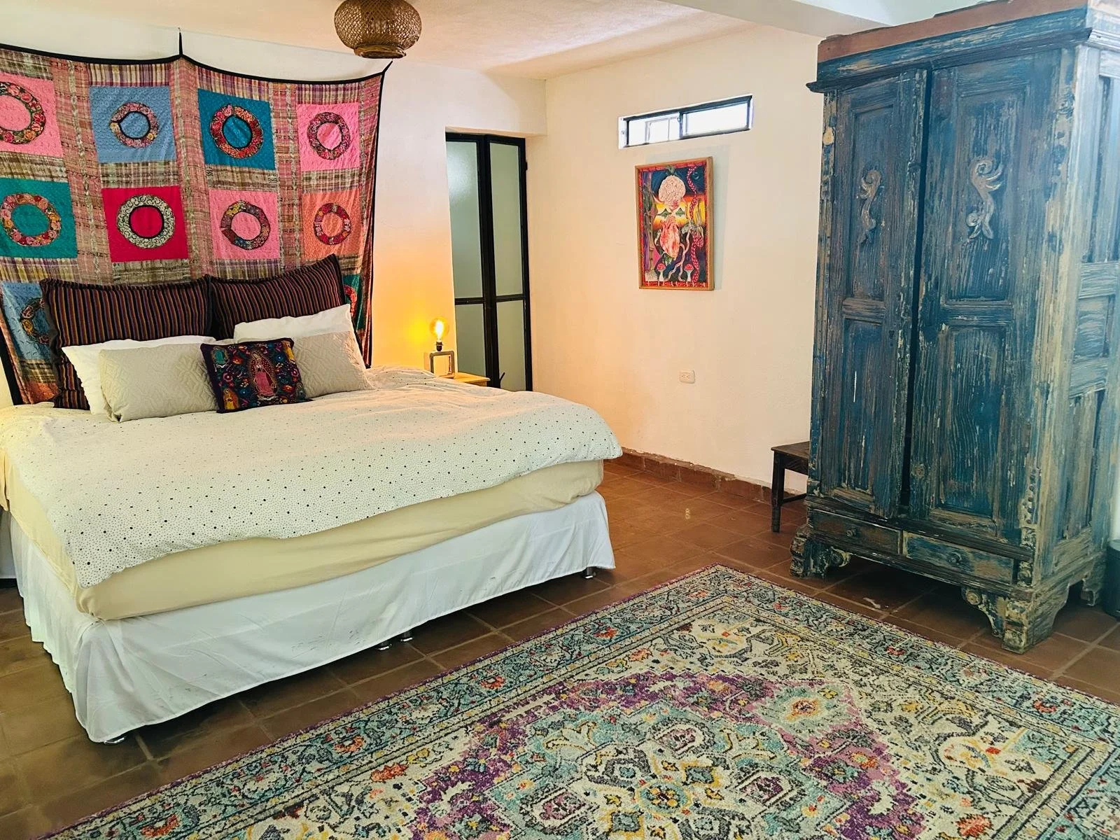 A bedroom featuring a bed with a colorful, patterned headboard, multiple pillows, and a white bedspread. There is a vintage blue-green armoire, a small side table with a lamp, and a decorative rug on brown tile flooring. Artwork hangs on the wall, an