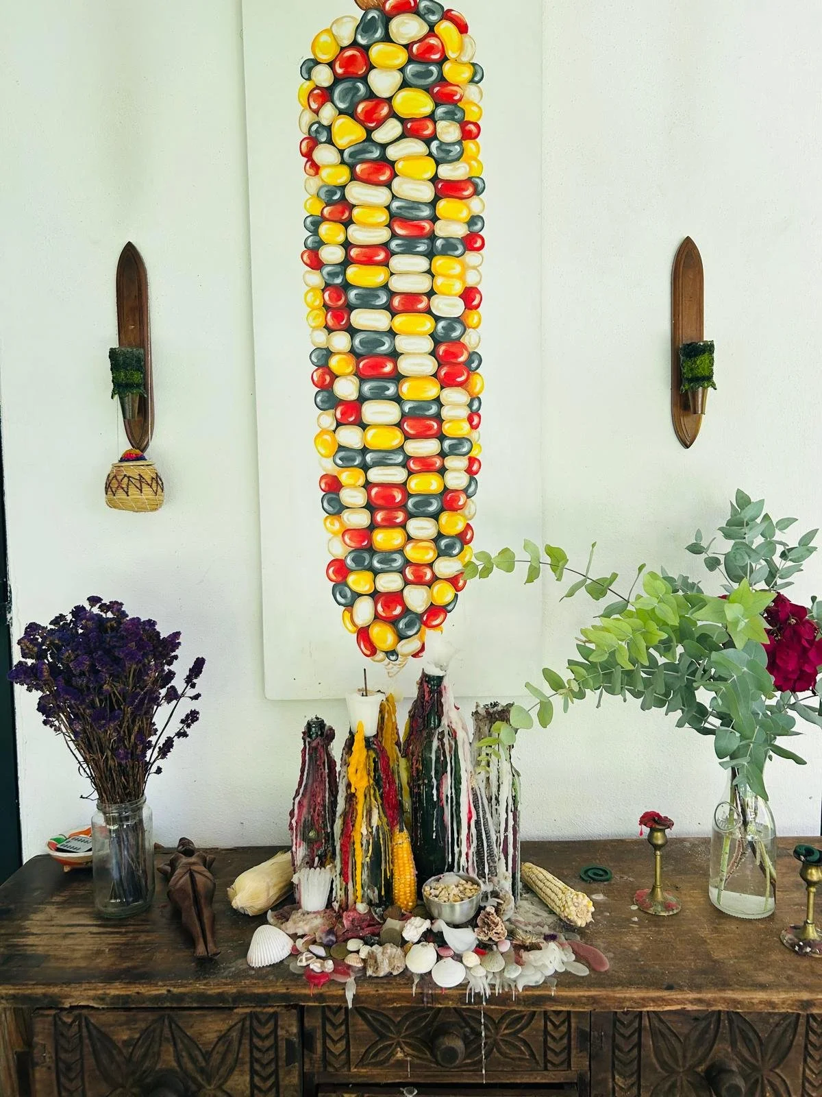 Decorative display with a vertical artwork made of colorful beans or stones, candles, seashells, dried flowers, and plants on a wooden table in front of a white wall.