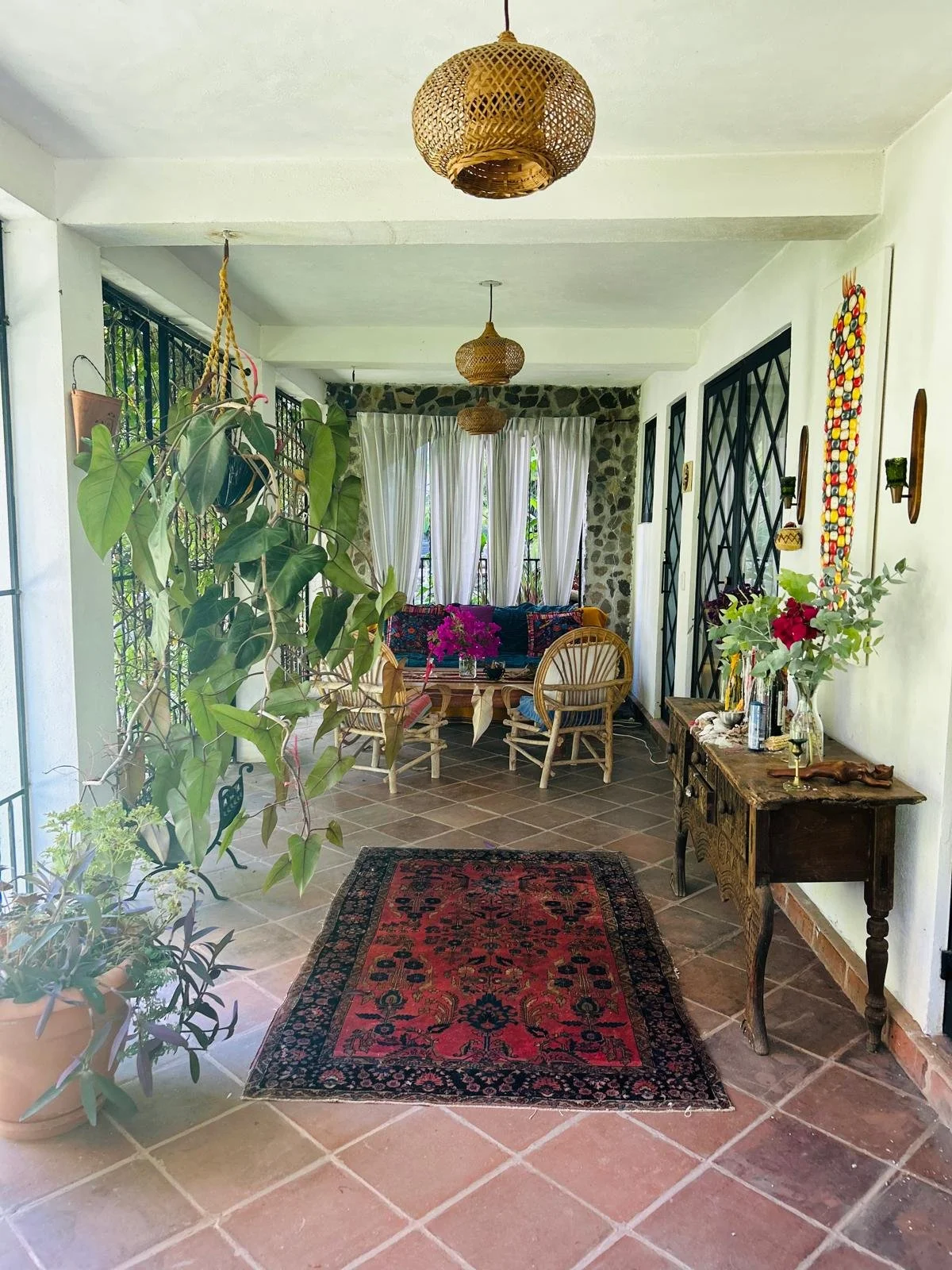 Indoor patio with potted plants, a red patterned rug, a wooden table, chairs, and hanging woven light fixtures.