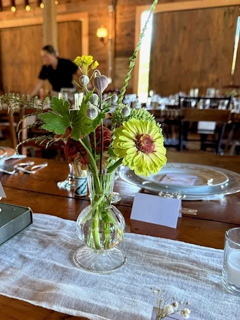 A glass vase with yellow and green flowers and greenery on a table indoors, with a blurry background of a wooden room and a person in black.