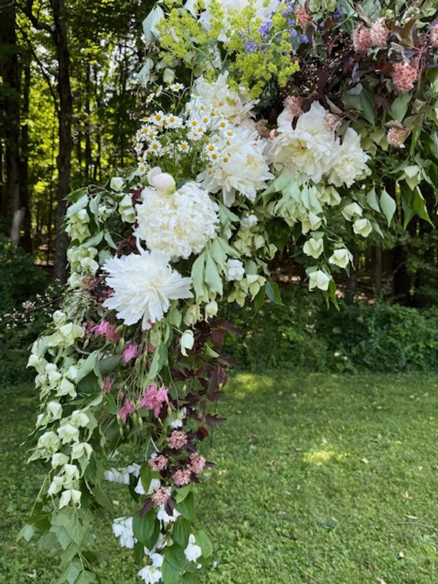 A floral arrangement with white peonies, daisies, pink and purple flowers, and green foliage set outdoors with trees and grass in the background.