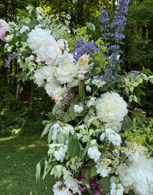 A lush floral arrangement featuring white peonies, hydrangeas, and daisies with purple and pink accents, set outdoors with a green forest background.