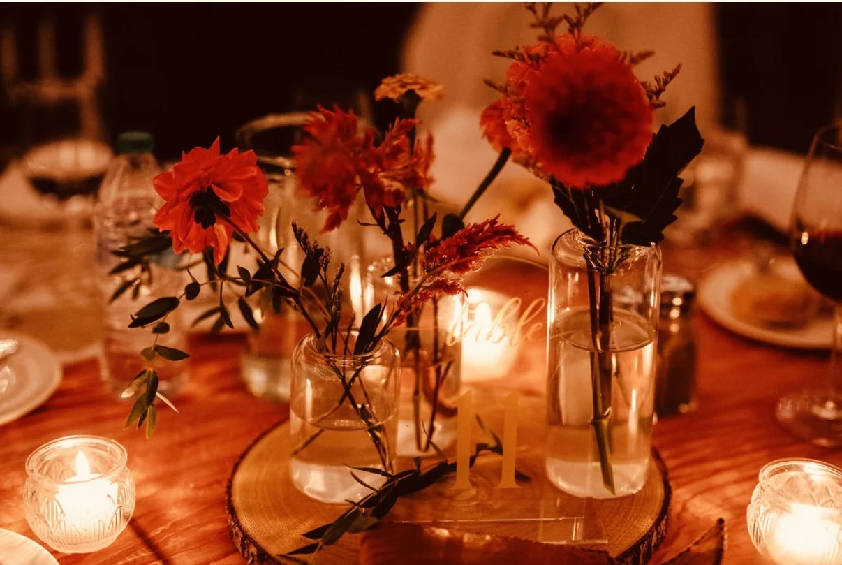 Table centerpiece with red flowers in glass vases, candles, and a wooden slice at a dinner event with warm lighting.