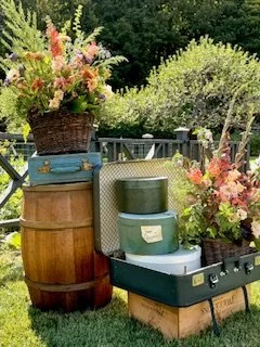 A vintage style picnic setup outdoors with a small green stove inside an open suitcase, surrounded by flower bouquets in baskets and containers, on a grassy lawn with trees in the background.
