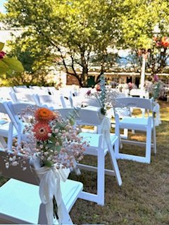 White chairs decorated with pink and white flower bouquets on a grassy outdoor setting, with trees and a blue sky in the background.