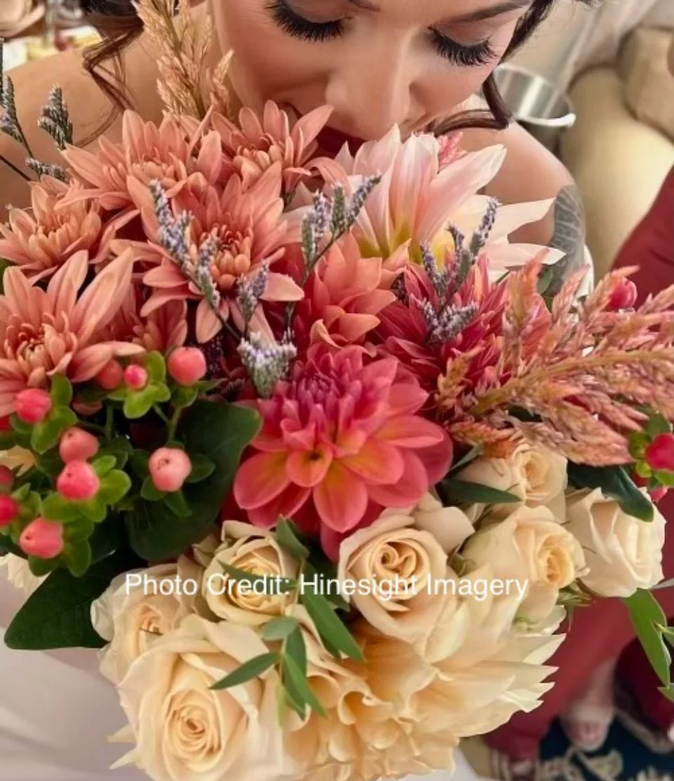 A woman is smelling a large bouquet of pink and peach flowers, including roses and dahlias, with some lavender and greenery.