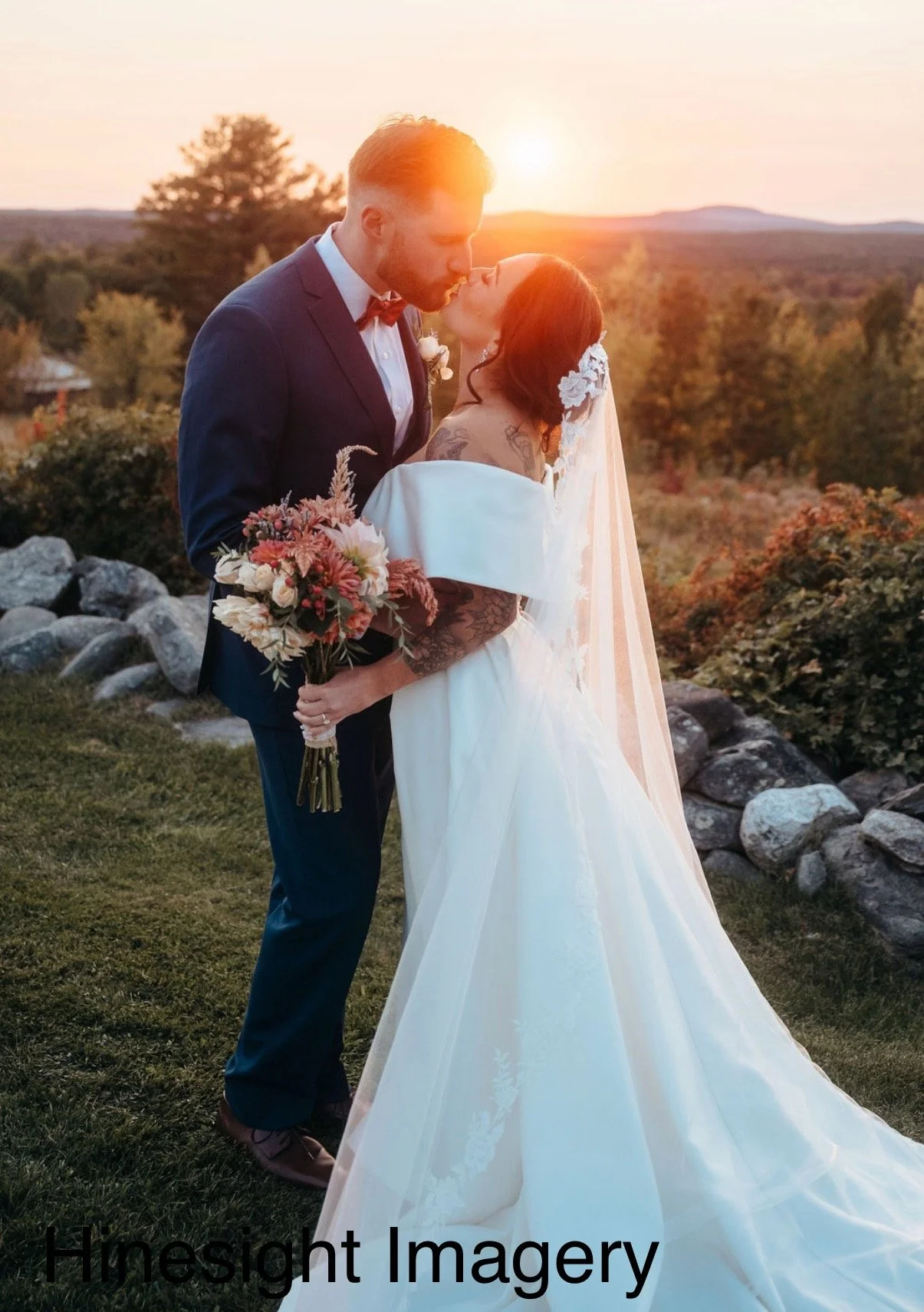A bride and groom sharing a kiss outdoors during sunset, with the bride holding a bouquet of flowers and wearing a white wedding dress with lace details, while the groom is dressed in a dark suit with a bow tie.