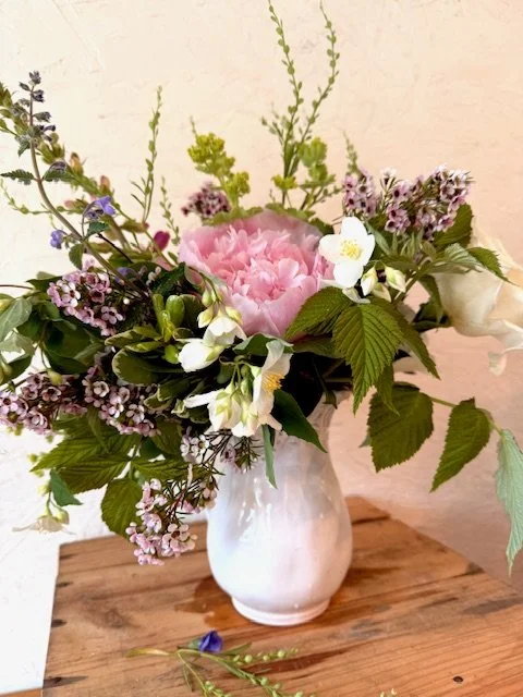 A bouquet of pink peonies, white flowers, and purple and green foliage in a white vase on a wooden surface.