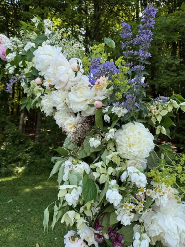 A tall floral arrangement with white, purple, and yellow flowers and green foliage outdoors in a wooded area.