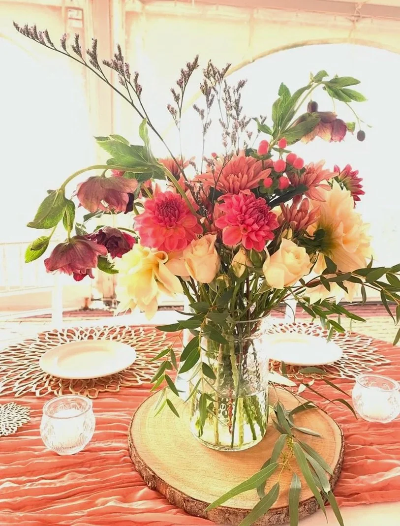 Flowers arranged in a glass vase on a wooden slice, placed on a pink tablecloth, with candle holders and plates on a decorated table.