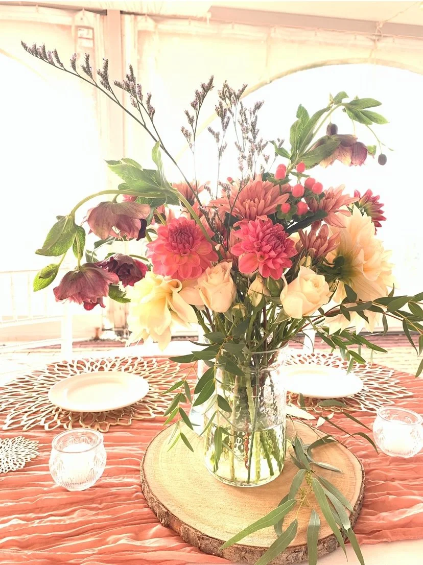 A dining table with a centerpiece of pink and cream flowers in a glass vase, placed on a wooden round slab. The table has coral-colored placemats, white plates, and small glass candle holders. The background is bright with a screened-in porch with curved openings.