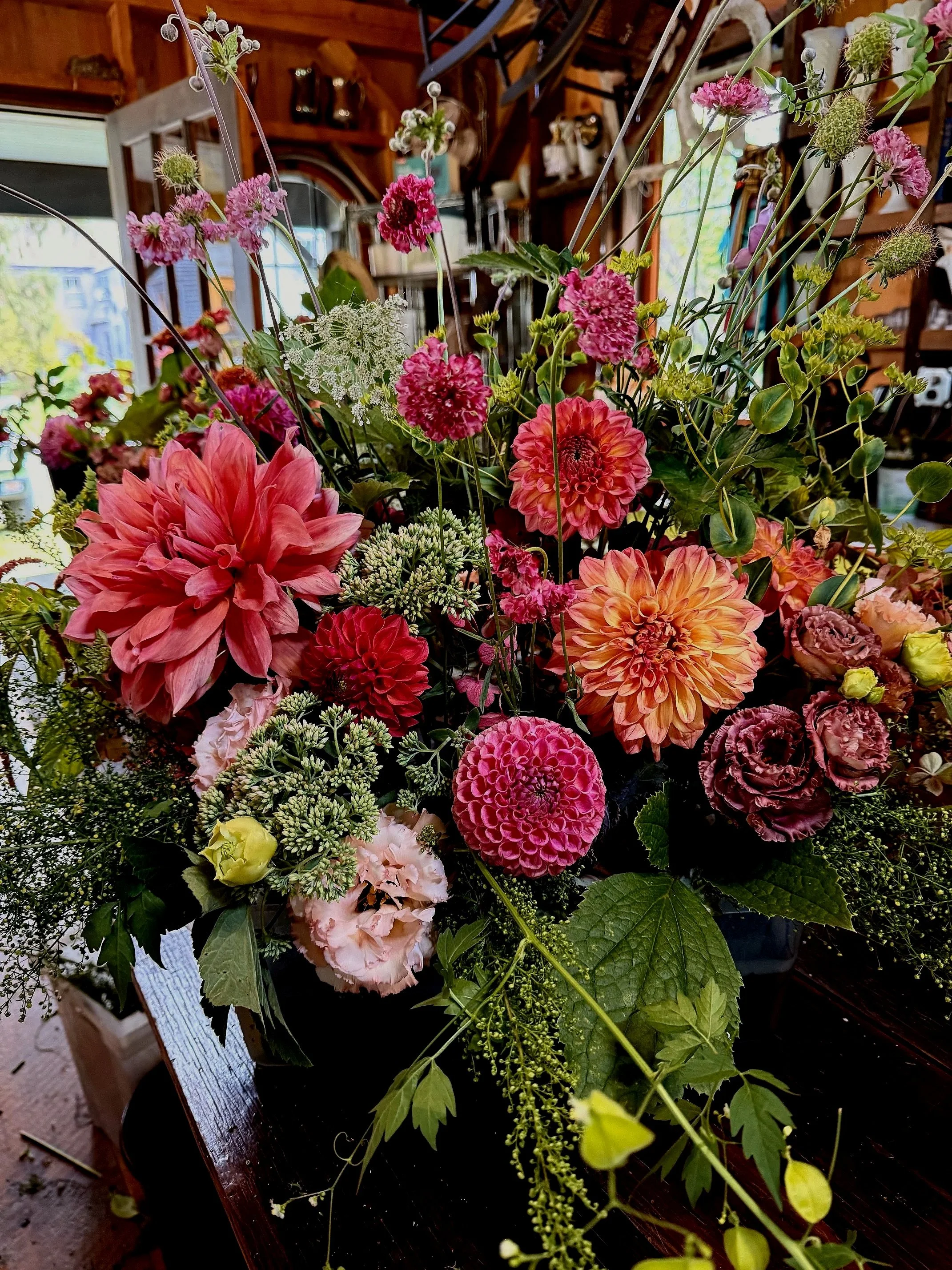 A colorful flower arrangement with pink, red, orange, and white flowers, including dahlias, zinnias, and other mixed blooms, set in a dark container on a wooden surface inside a cozy, rustic room.