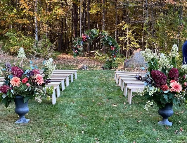 Outdoor wedding setup with flower arrangements and a floral arch in a wooded area.
