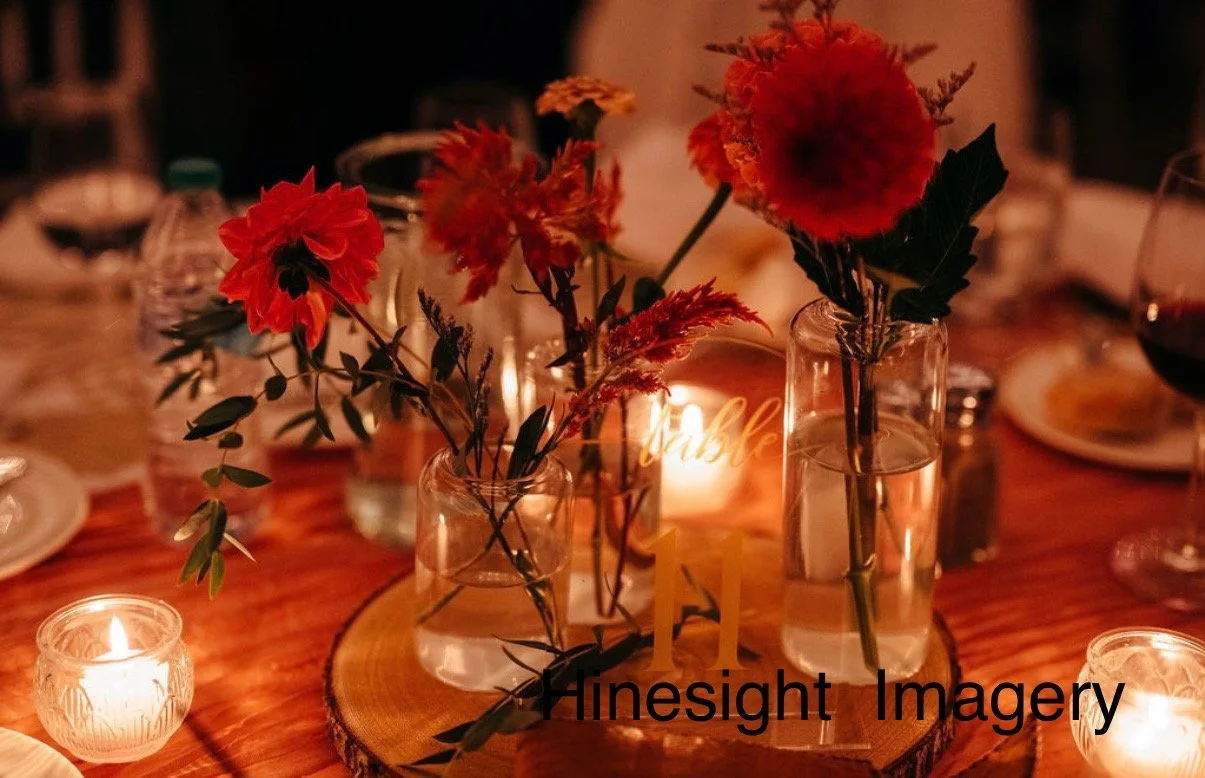 Decorative table centerpiece with red and pink flowers in glass vases, surrounded by lit candles on a wooden table at a warm, intimate event.