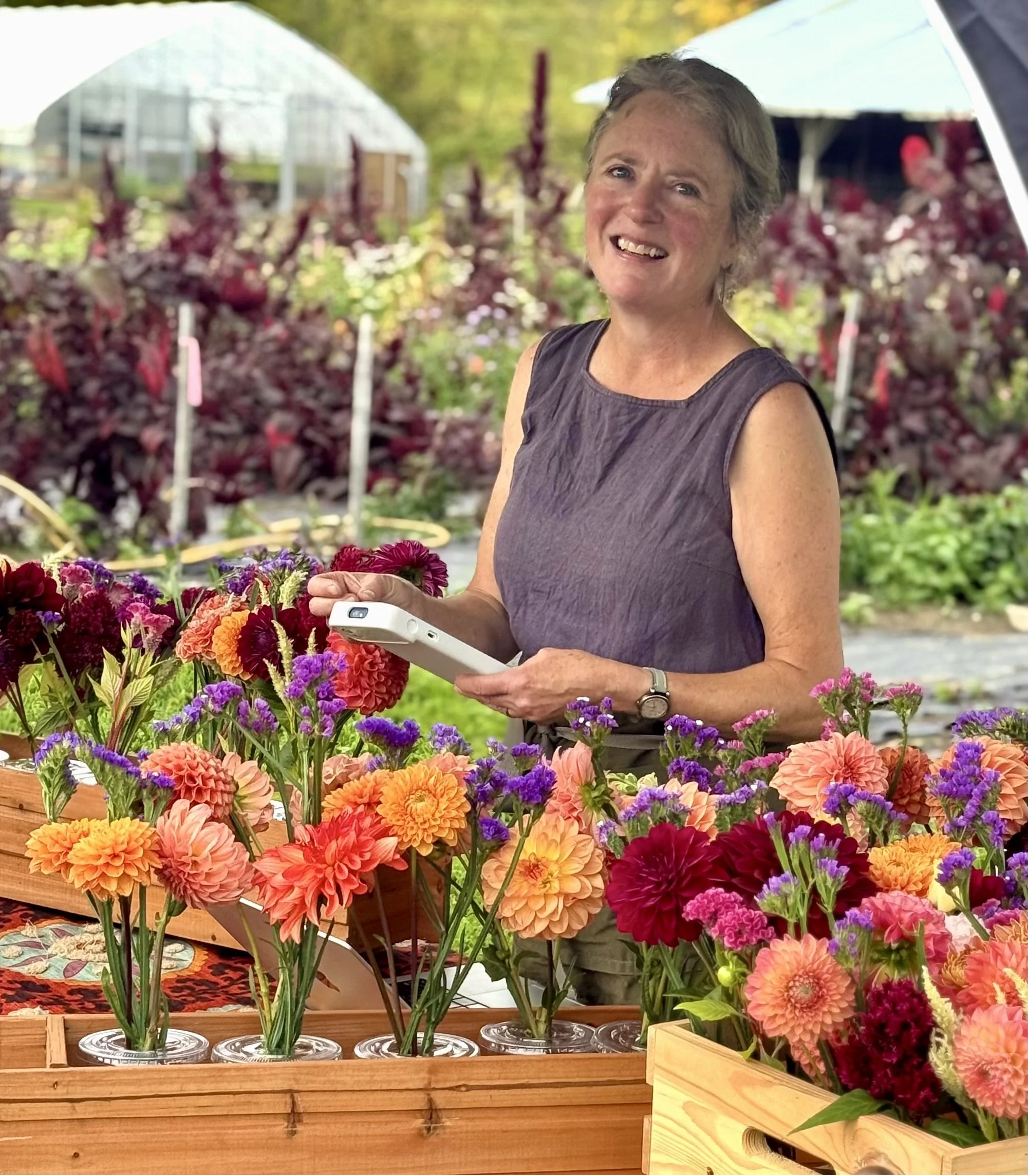 A woman stands behind a table of colorful flowers at a flower market, holding a white device, smiling with a garden and greenhouse visible in the background.
