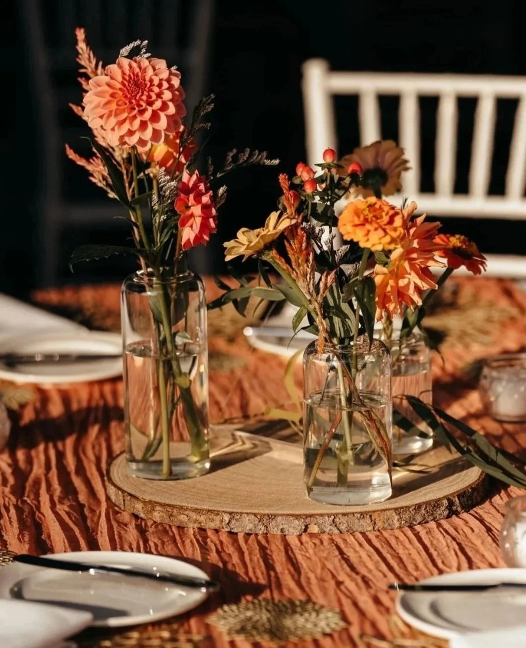 Vase with orange and red flowers on a wooden round base on a textured orange tablecloth, with plates and utensils nearby, in a dining setting.
