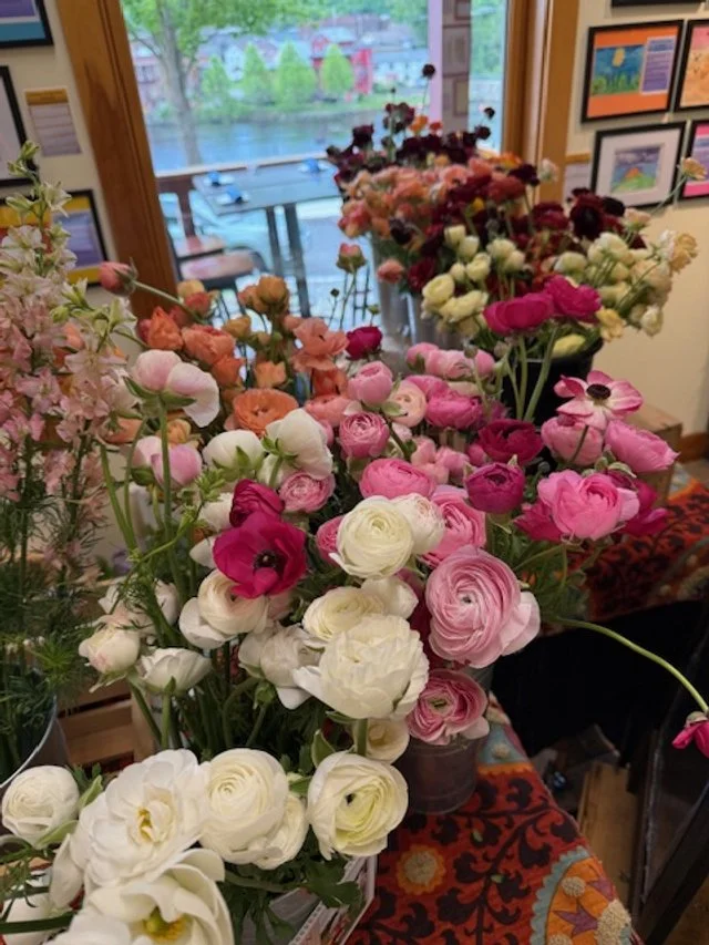 Various colorful flowers arranged on a table inside a room with artwork on the walls and a window showing trees and buildings outside.