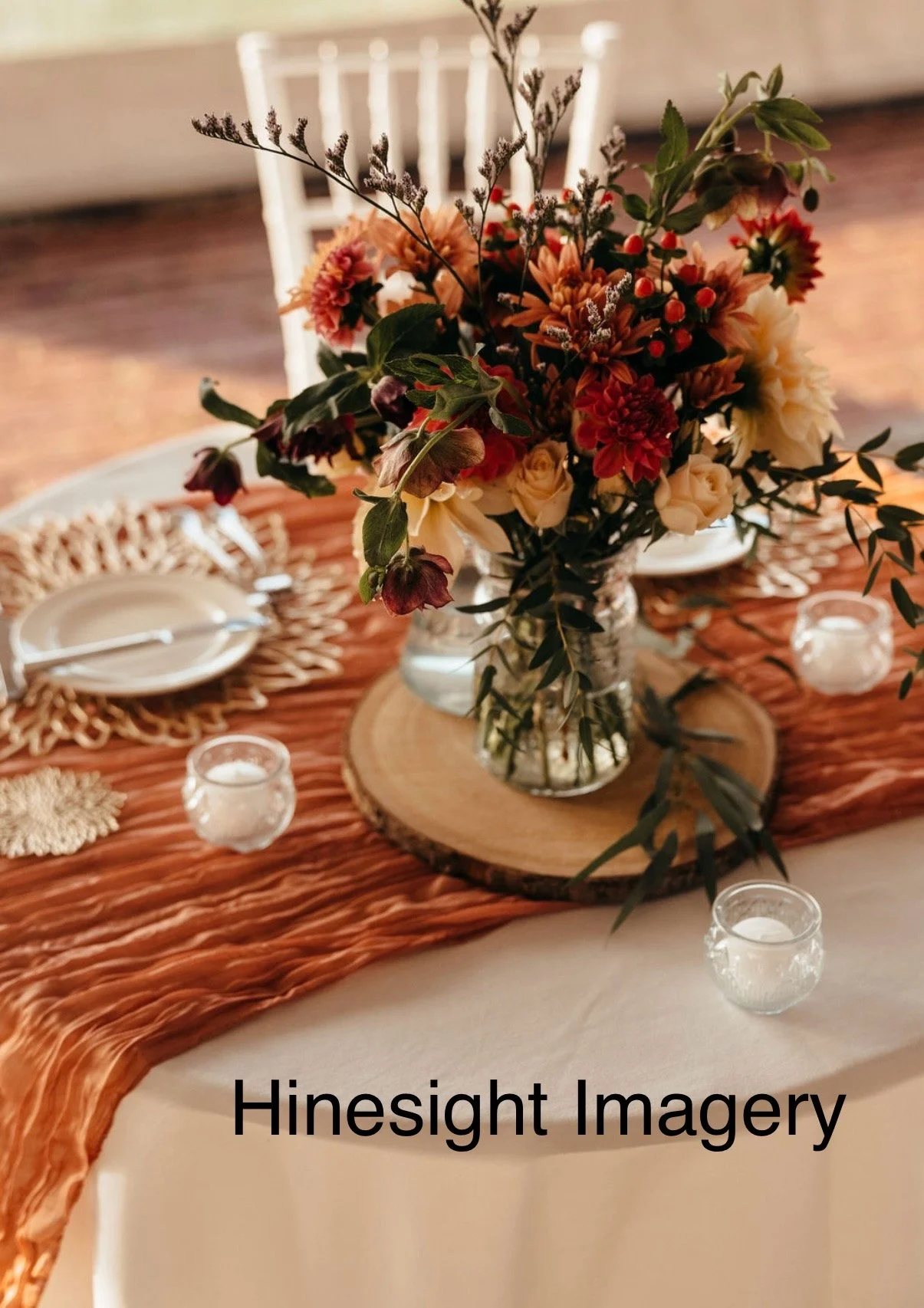 A floral centerpiece with mixed flowers and greenery in a glass vase on a wooden slab, surrounded by small candles, on a table with a rust-colored runner and white tablecloth.