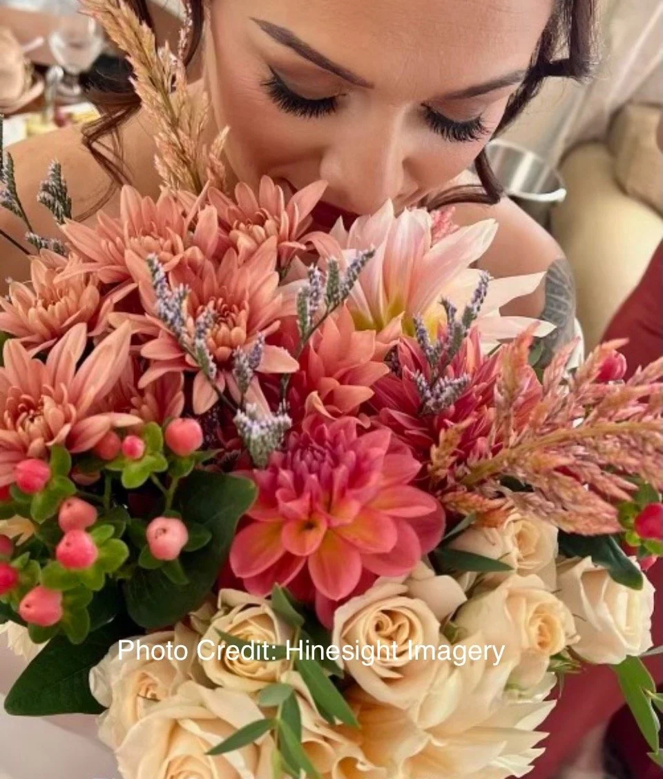 A woman with dark hair, closed eyes, and long eyelashes smelling a vibrant bouquet of pink, cream, and purple flowers.