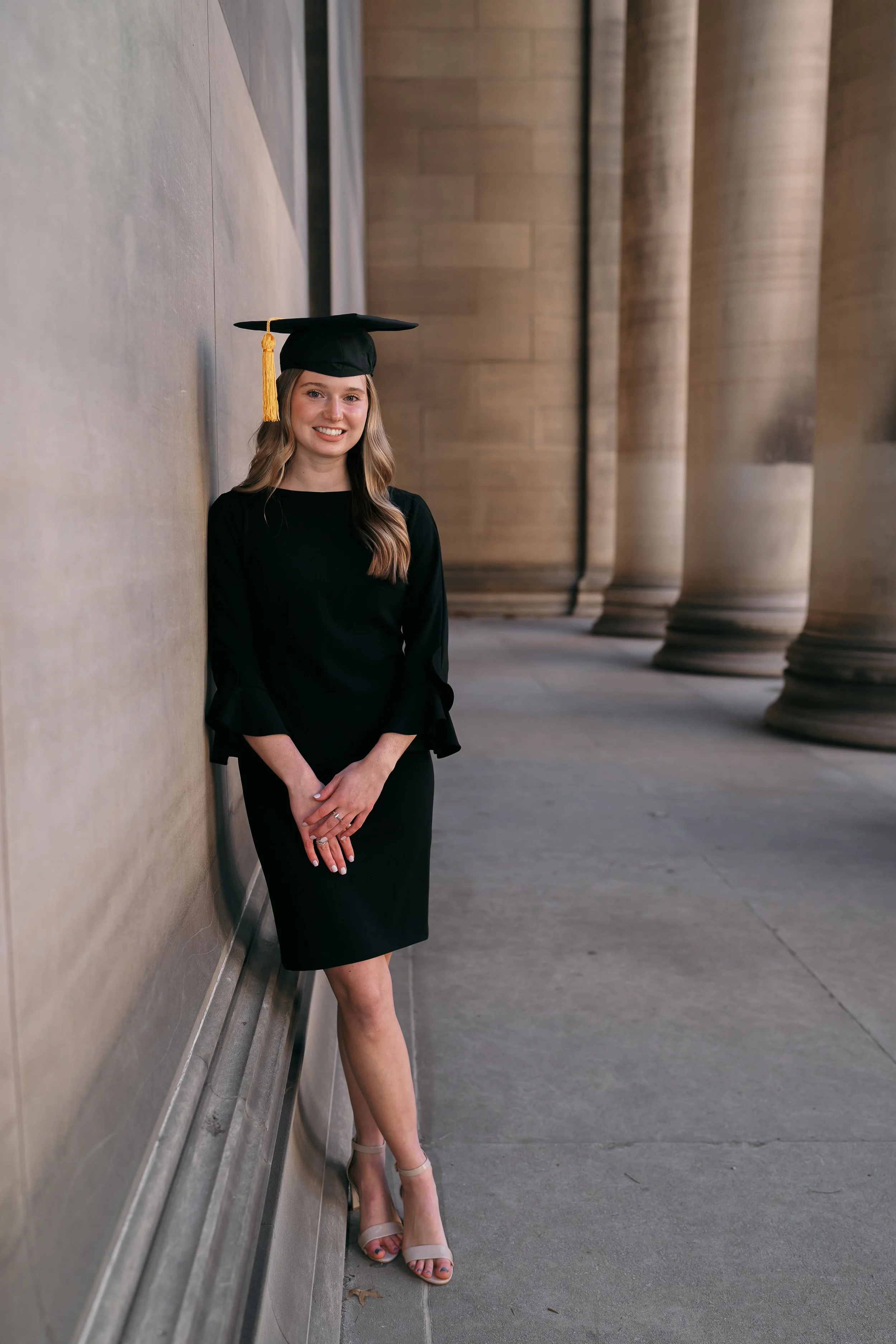 A young woman in a black dress and high heels wearing a graduation cap with a gold tassel leaning against a wall with large columns in the background.