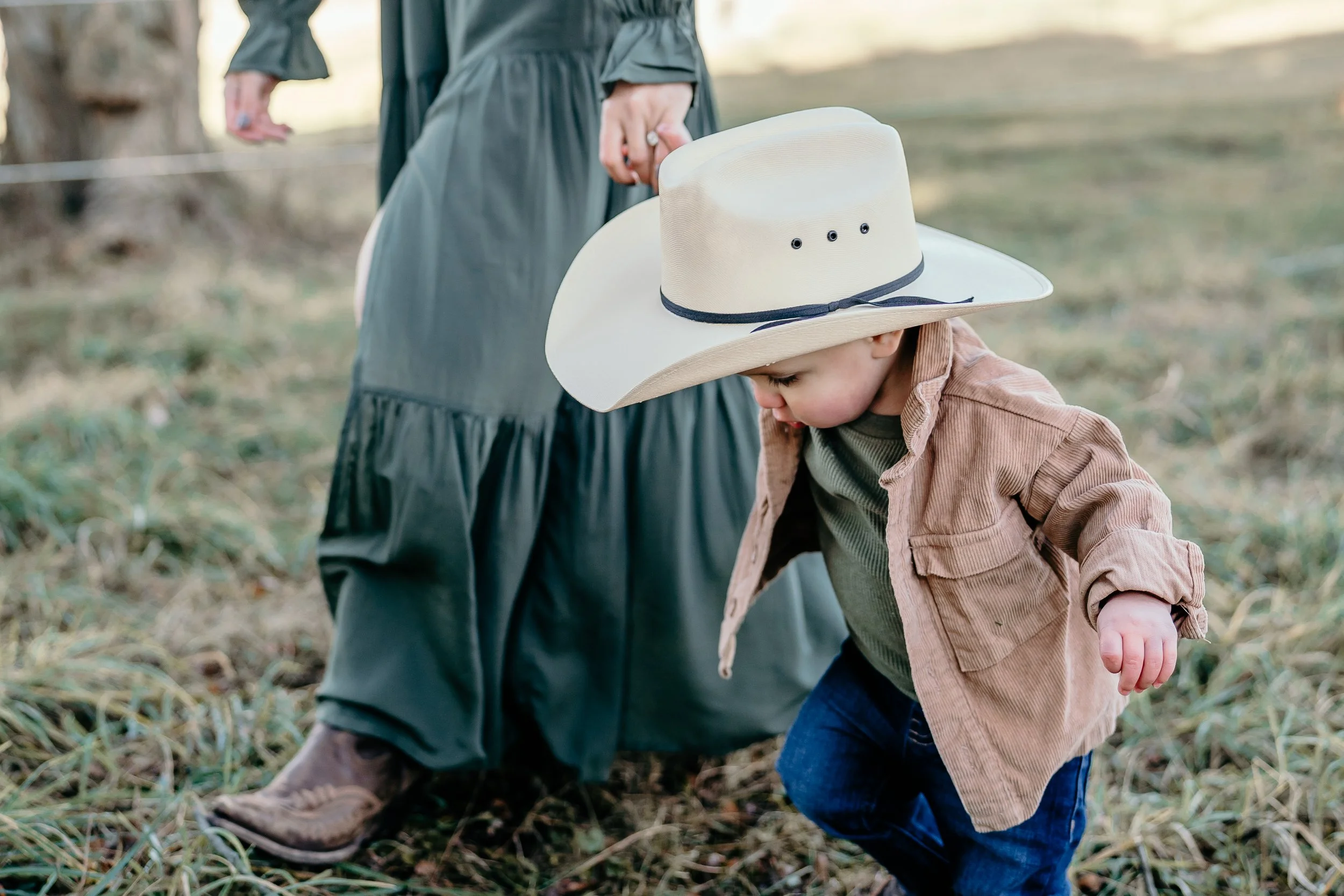 A young boy with a large cowboy hat, brown corduroy shirt, green T-shirt, and jeans is crouching and looking down at the ground in a grassy outdoor area. An adult wearing a long dark dress and boots is standing nearby.