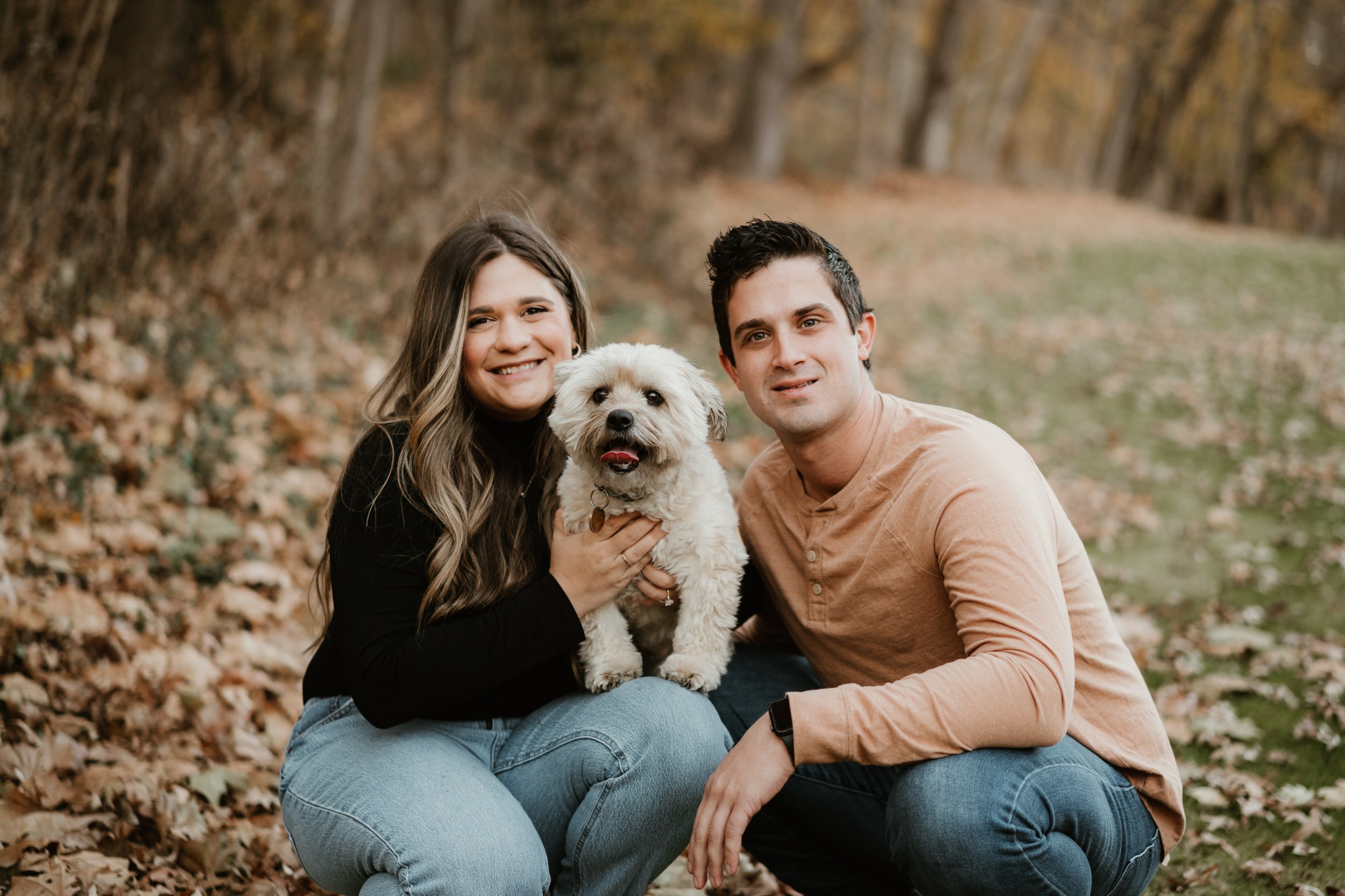 A smiling woman and a man are outdoors in a park, holding a small fluffy dog between them, with fallen autumn leaves on the ground.