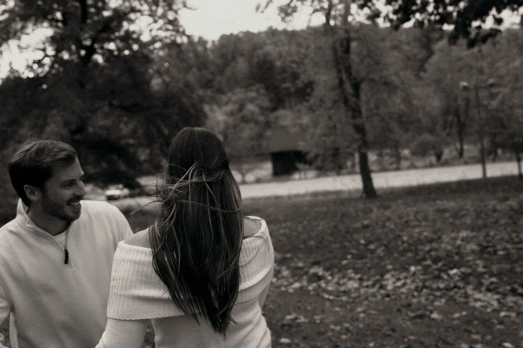 A man and a woman smiling at each other outdoors in a park with trees and fallen leaves.