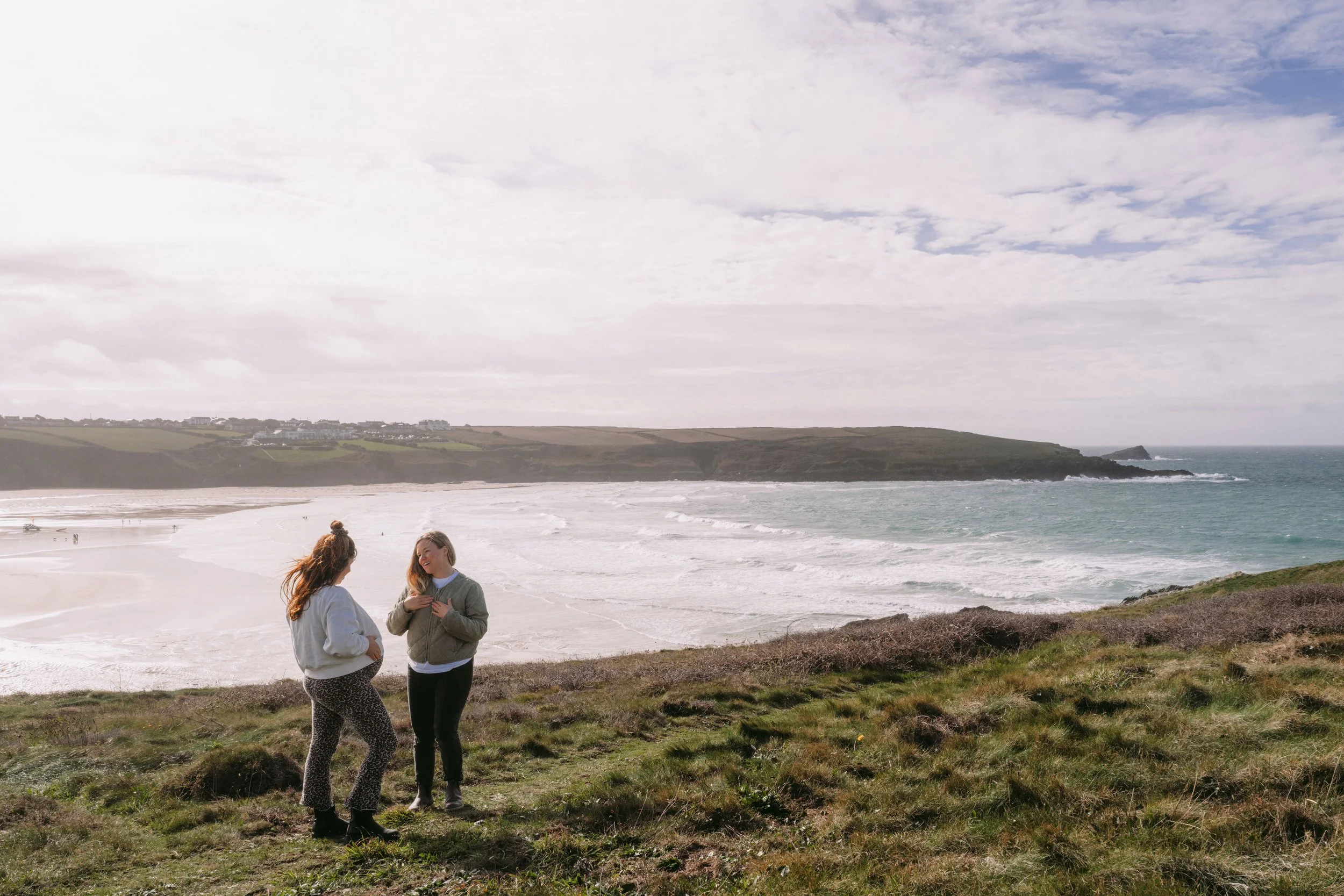 Pregnant cornish woman stands on a cliff with her personal private midwife. The background shows rolling waves, the Newquay pentire headland and a blue sky with clouds. They are discussing pregnany massage, homebirth and breastfeeding