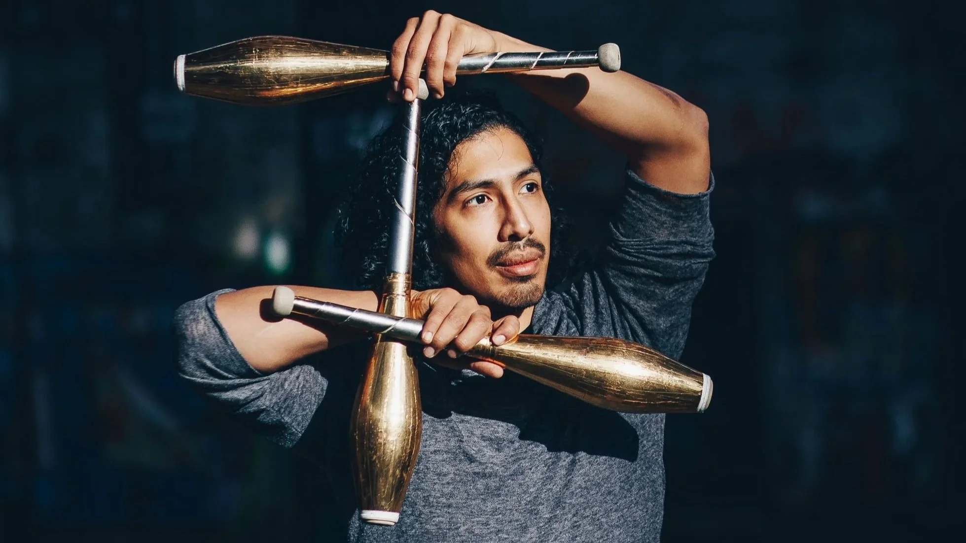 Ein Mann (Fabio Zimmermann) mit langen, lockigen Haaren jongliert mit drei goldenen Keulen in einem dunklen Raum.