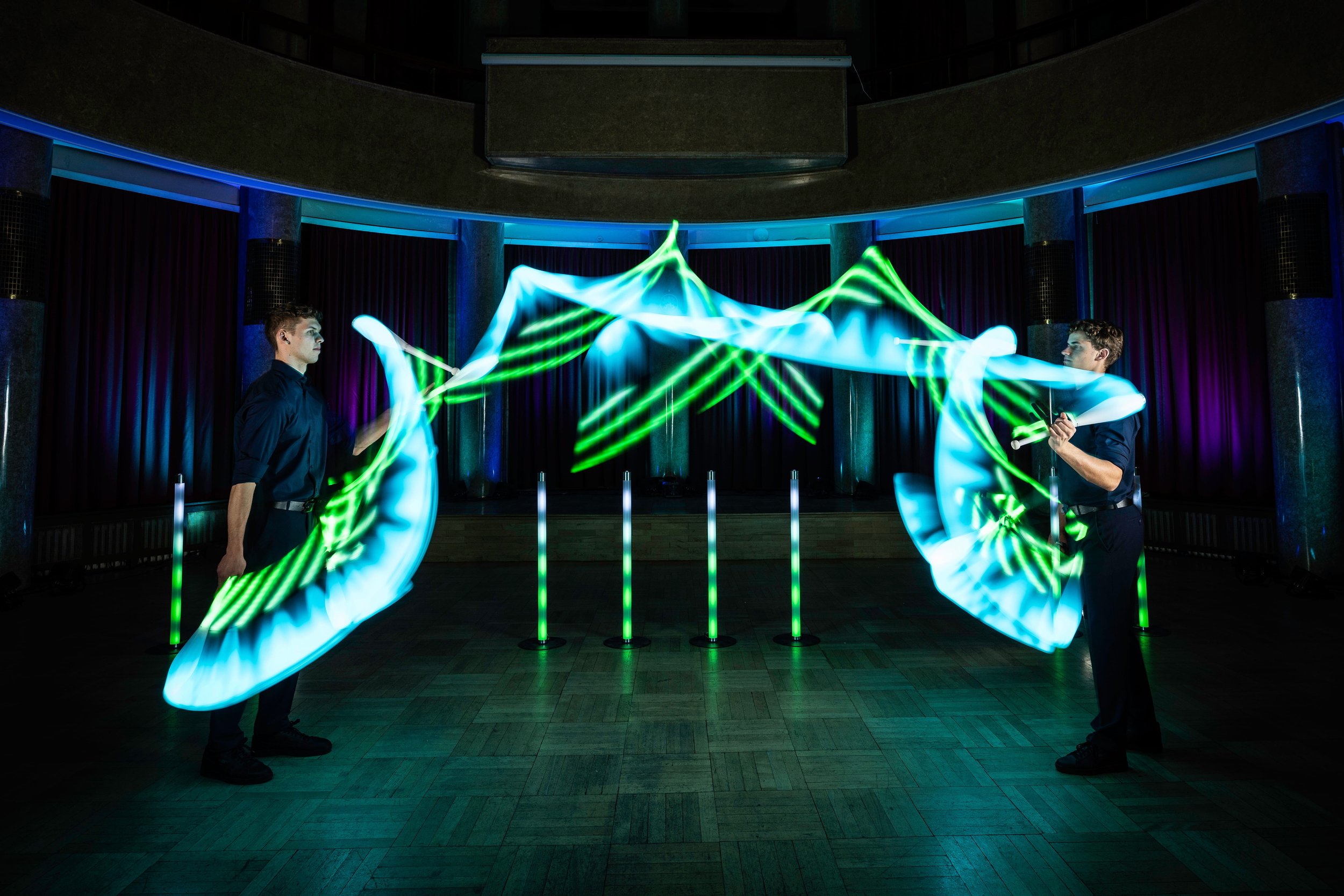 Zwei Personen führen eine Lichtshow mit leuchtenden Stäben in einem abgedunkelten Raum auf, umgeben von säulenartigen Strukturen. Leuchtende Linien in Blau und Grün erzeugen dynamische Muster.