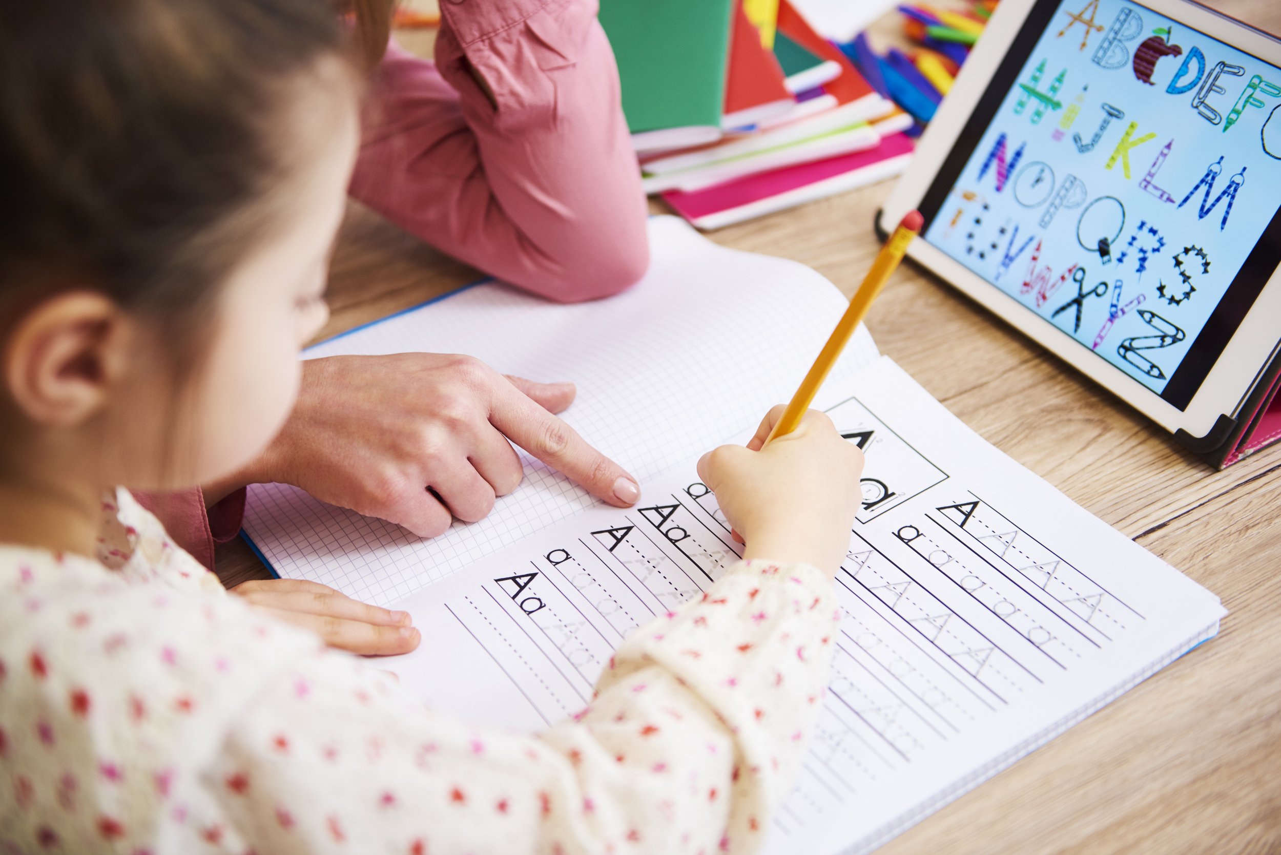 Young child practicing handwriting with a pencil on a notebook, guided by an adult pointing at letters. An alphabet chart is displayed on a tablet nearby.