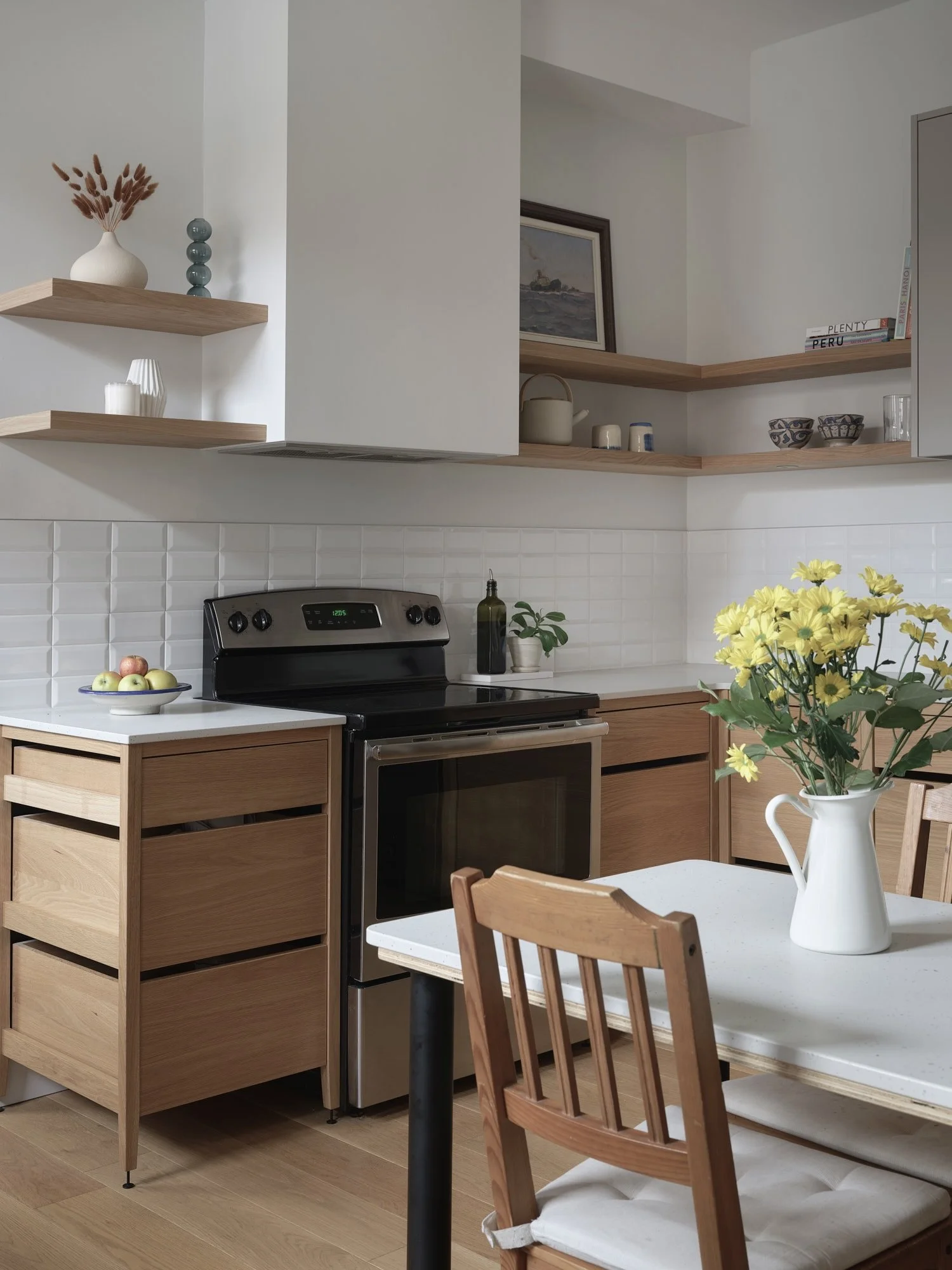 Cuisine modulaire sur pattes en chêne naturel, comptoirs blancs, îlot servant de table, chaises en bois et bouquet jaune. Électroménagers en inox, étagères flottantes en chêne et dosseret en carreaux de métro blanc.