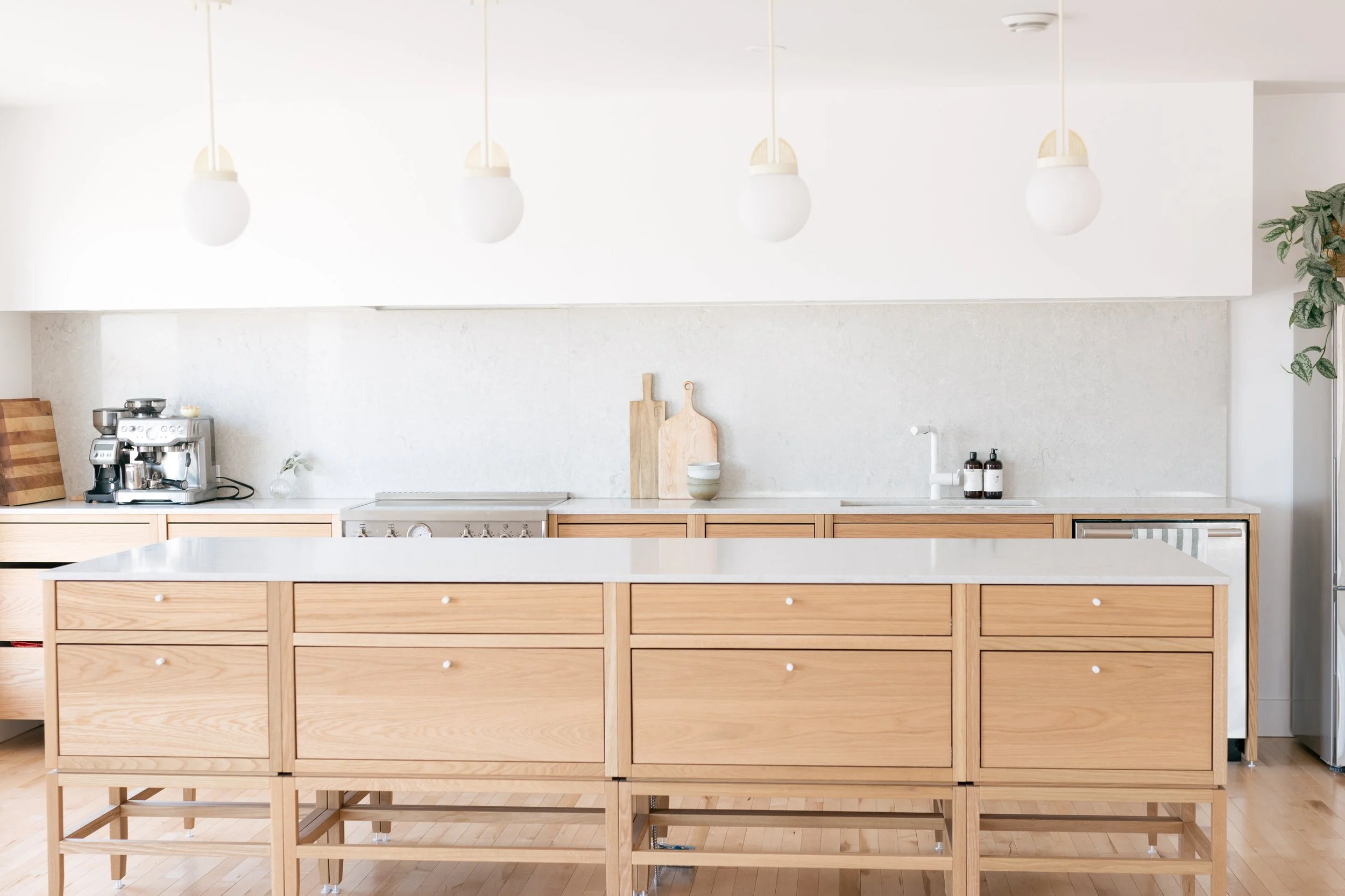 Cuisine moderne avec îlot en chêne naturel, comptoir en marbre blanc, suspendus en forme de demi-sphère blanche, quelques éléments décoratifs en bois et vaisselle sur le plan de travail.