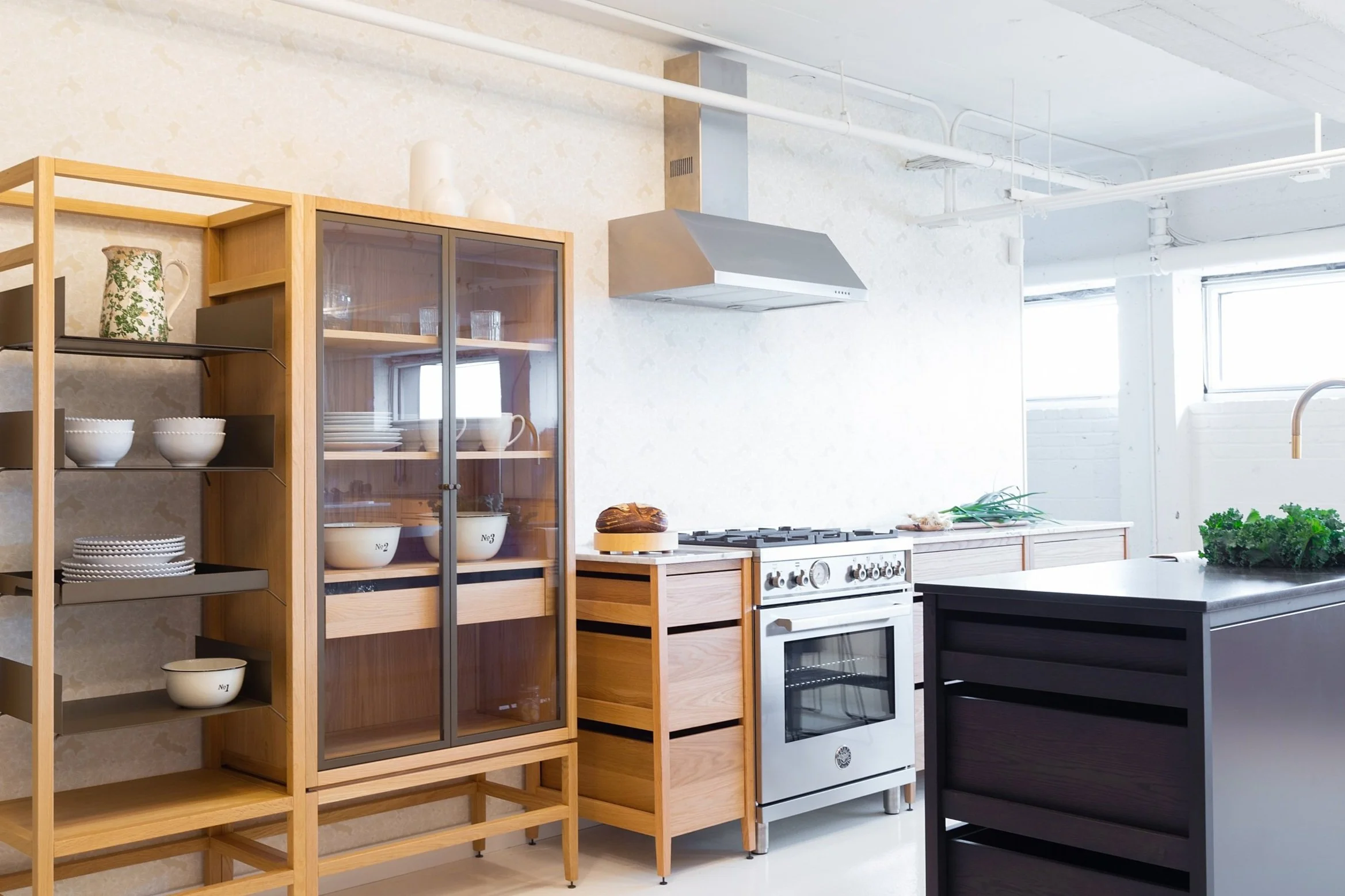 Cuisine avec armoires modulaires en chêne naturel, îlot en chêne teint noir, hotte en inox apparente, vaisselier vitré sur pattes et grande étagère en métal et bois.
