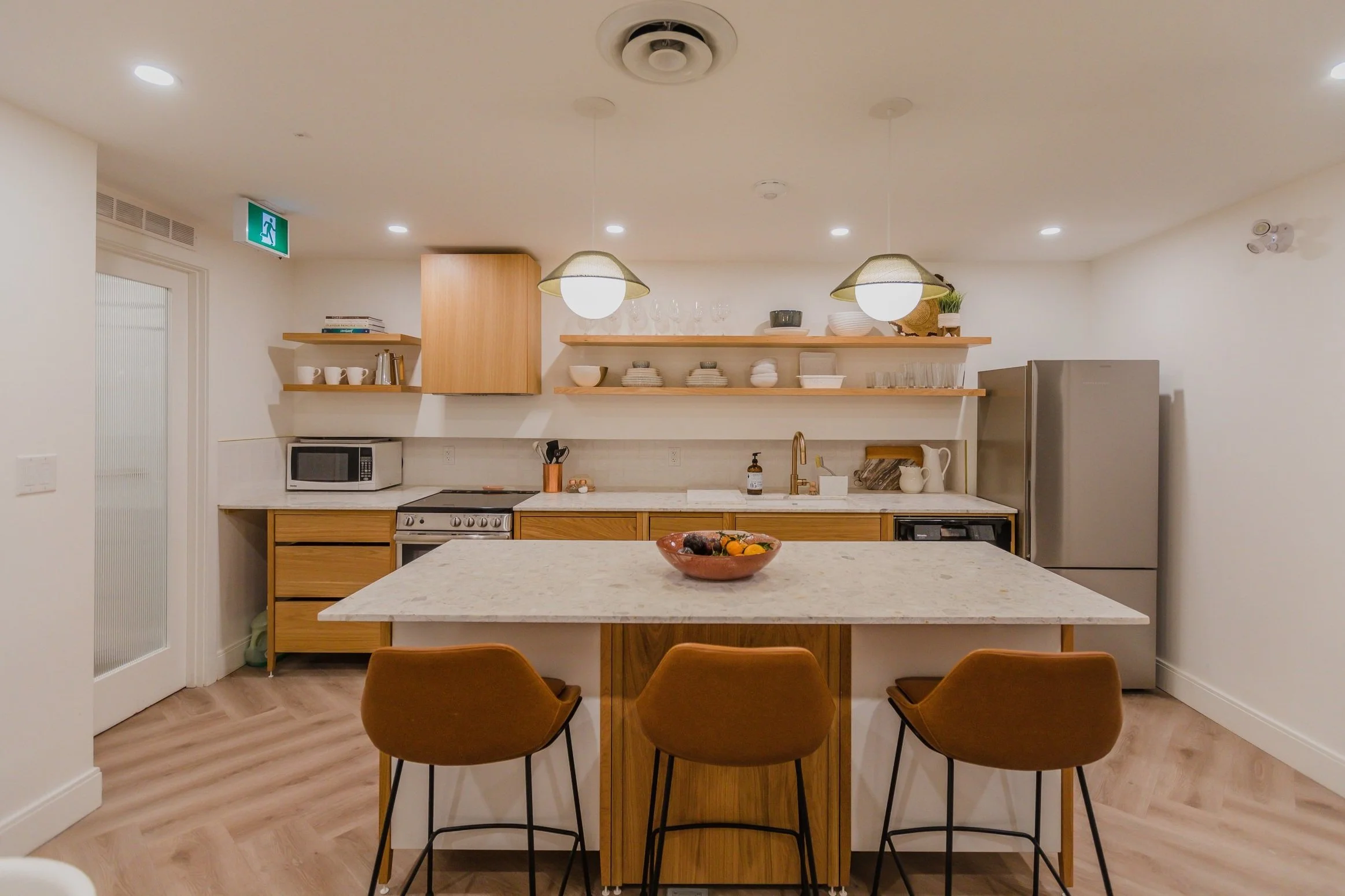 Cuisine moderne avec îlot central en marbre, chaises en bois brun, étagères en bois clair avec vaisselle blanche, réfrigérateur en acier inoxydable, four, micro-ondes, et éclairage suspendu