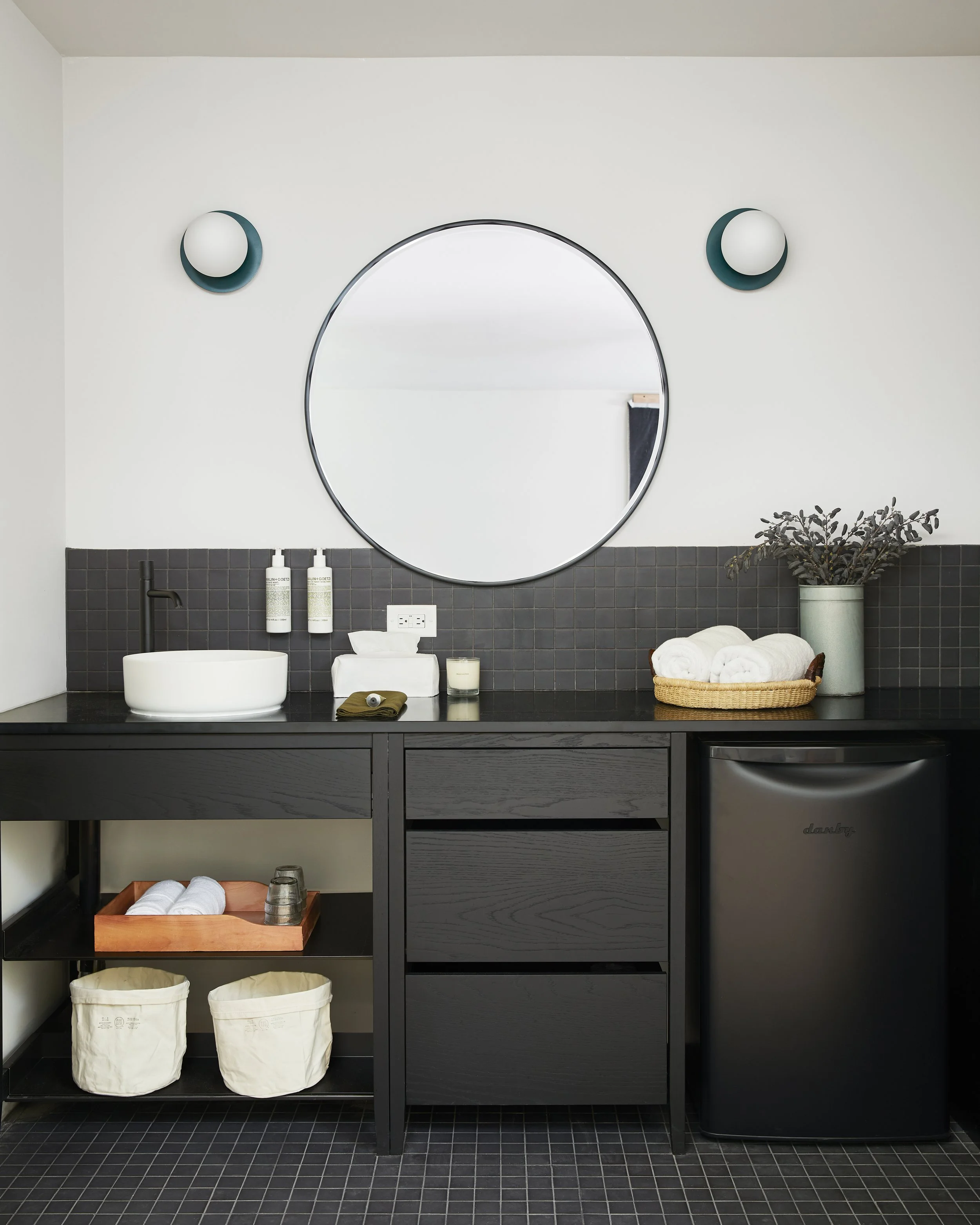 Une salle de bain moderne avec un miroir rond, un évier en bowl blanc, un comptoir noir, une petite cave à vin noire, et des accessoires comme des serviettes, des produits de bain, et une plante vert foncé.