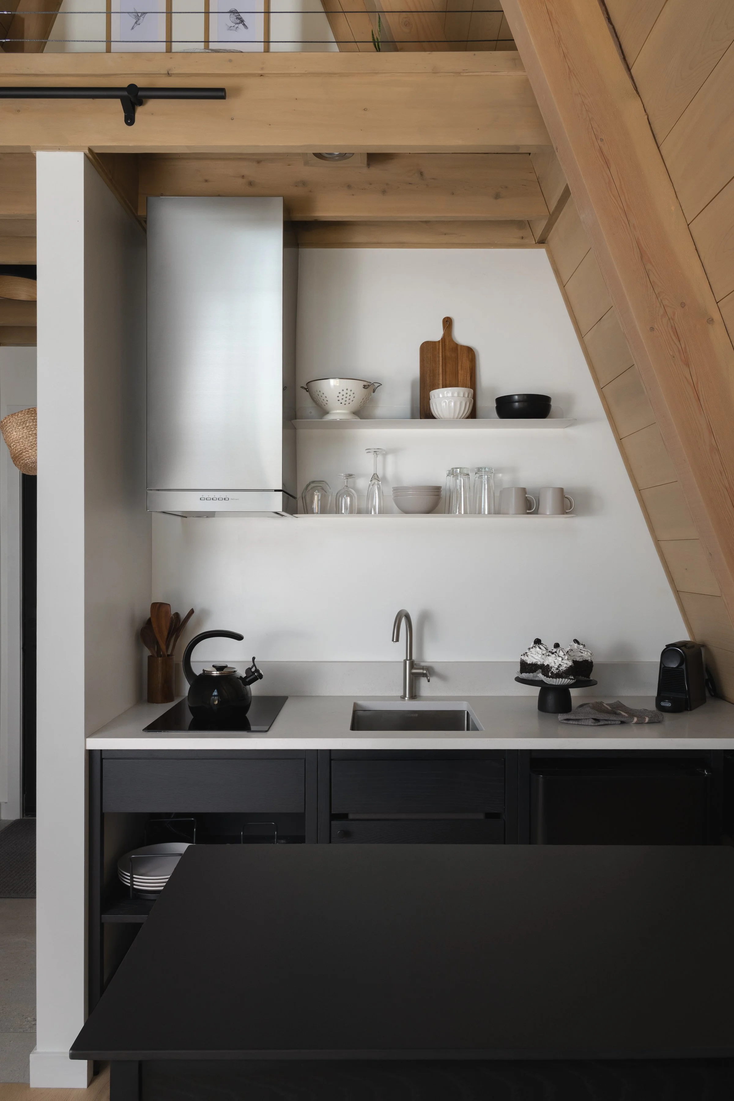 Cabine A-Frame, une design épuré, concept ouvert, avec une cuisine modulaire coquo en chêne teint noir et comptoir noir à l'îlot et comptoir et dosseret clair contre le mur arrière. Tablettes flottantes en métal blanc, avec hotte inox.
