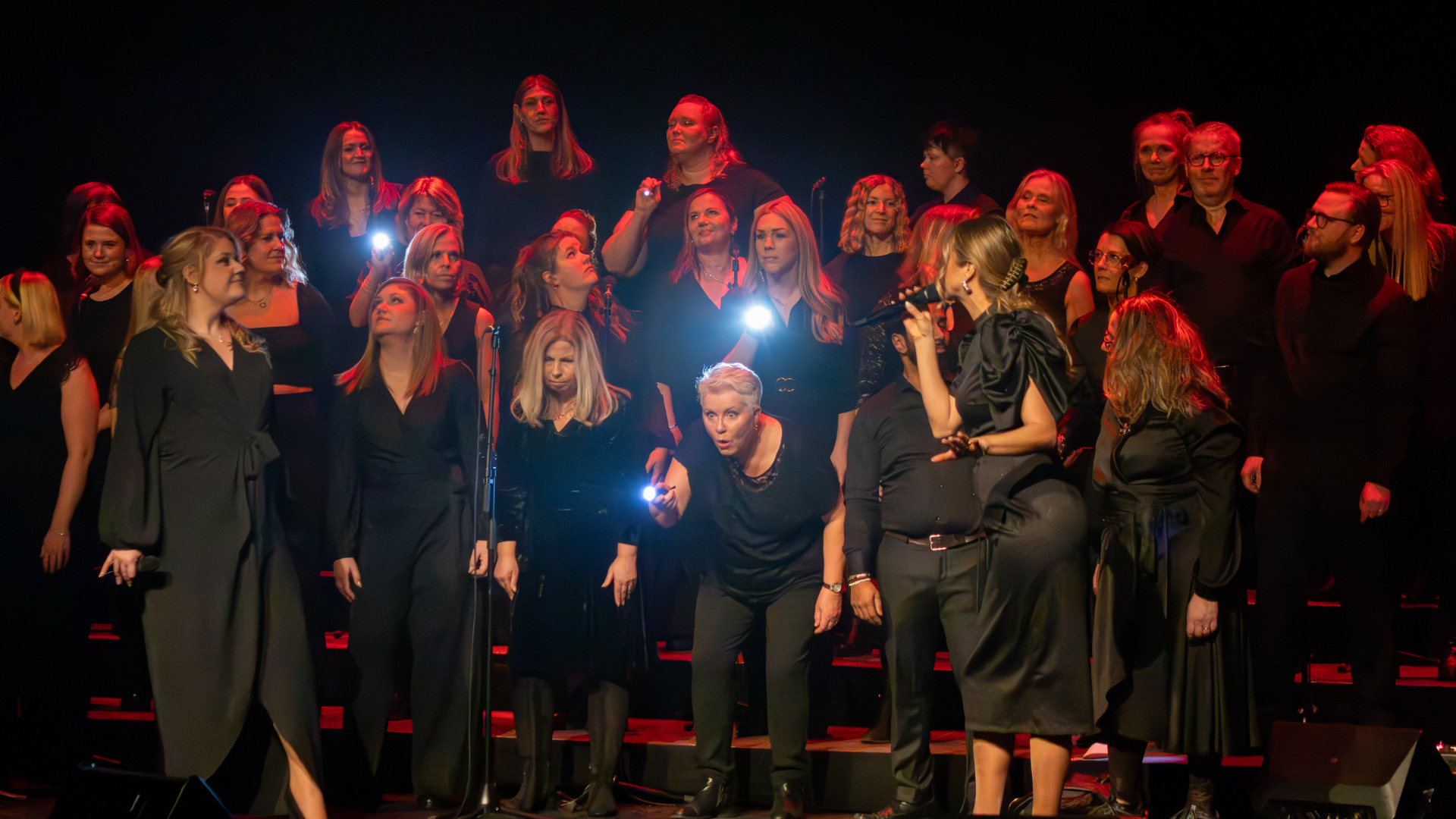 En grupp silverhåriga och unga kvinnliga och manliga sångare i svarta kläder, på scen, med två kvinnliga ledare framför, en med mikrofon och en med ficklampa, under en sånguppvisning.