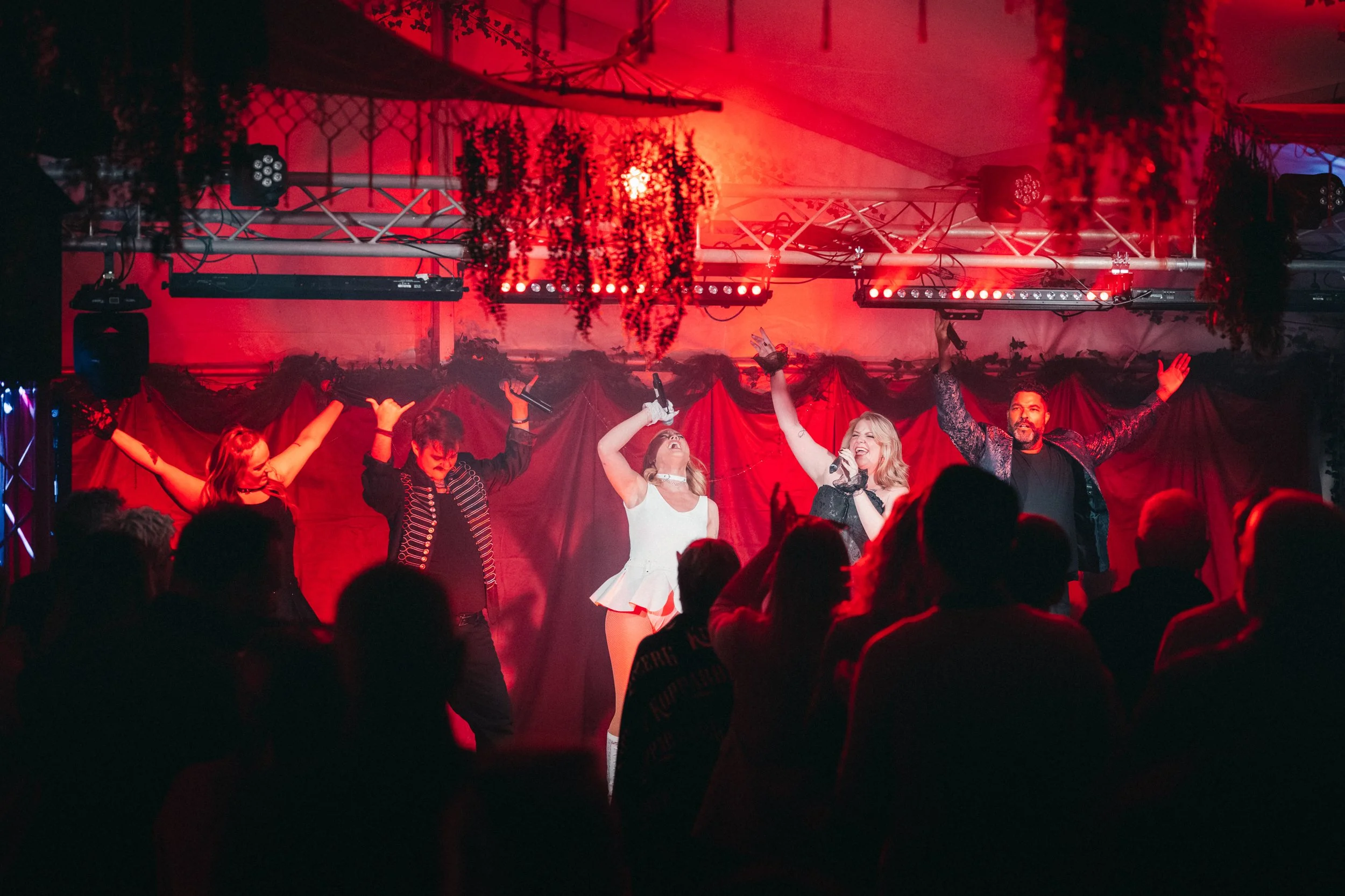Fem personer på scenen under en konsert eller show, med röda ljus och en publik framför.
