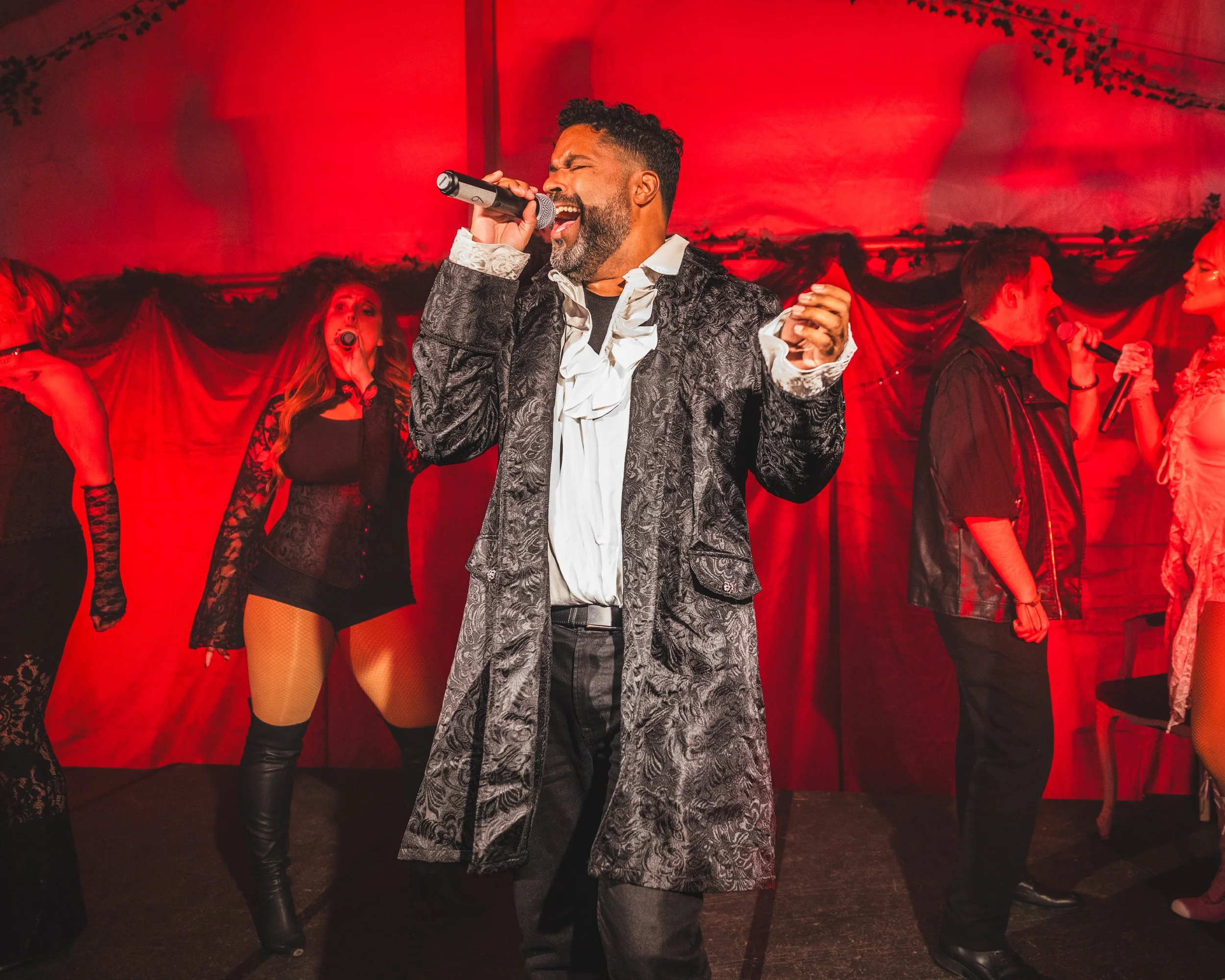 Grupp av personer som sjunger och dansar på en scen med röd bakgrund, ett manligt gospel- eller showkör med mikrofon i handen i centrum.