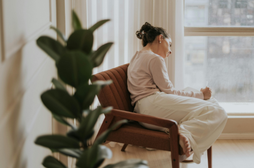 A woman sitting on a brown chair by a window, looking down with a thoughtful expression, with a large green plant in the foreground.