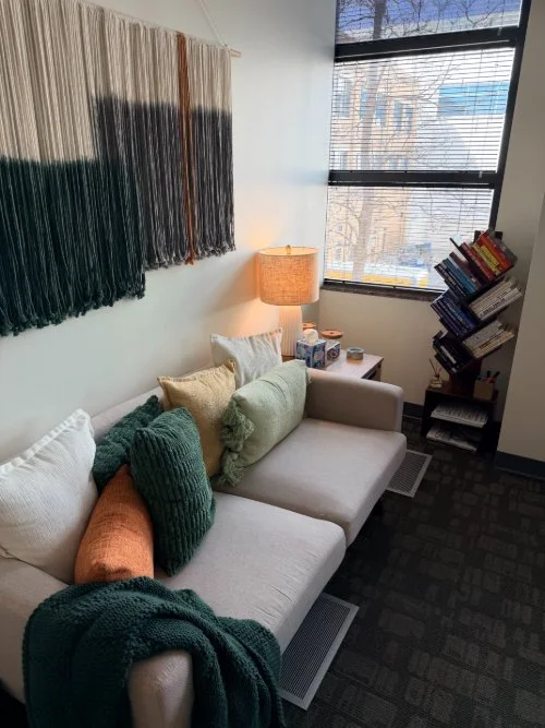 A cozy living room corner with a white sofa decorated with colorful cushions, a wooden side table with a lamp, a window with blinds, and a tilted bookshelf filled with books.