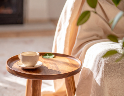 A beige coffee cup on a wooden tray next to a small green leaf with sunlight streaming in, near a beige sofa with a green plant in the background.